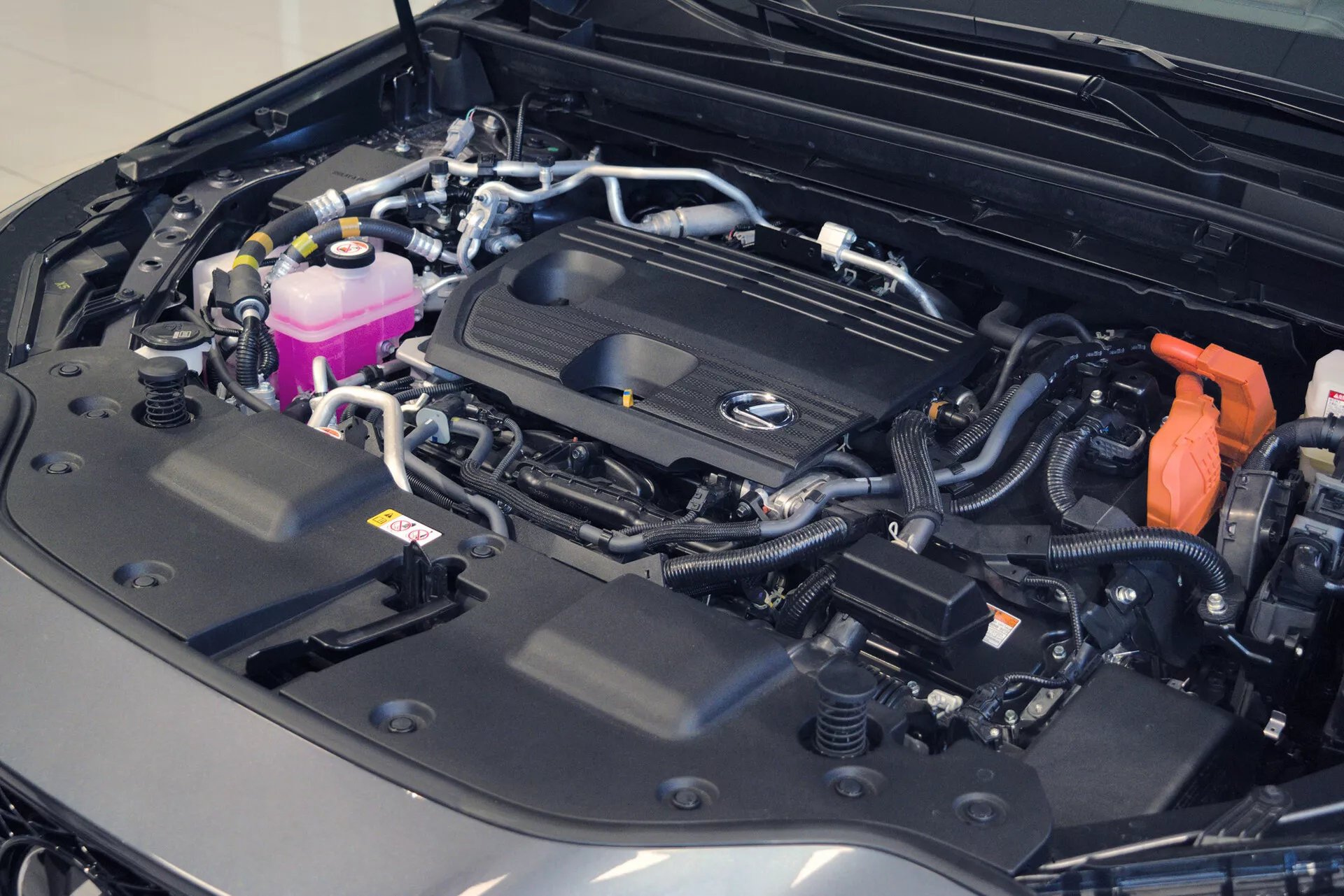 Detailný pohľad na motorový priestor auta s otvorenou kapotou, odhaľujúci dobre organizovaný a čistý motorový priestor. Kryt motora je opatrený výrazným logom, ktoré naznačuje značku, a rôznymi komponentmi, ako sú farebné nádrže, zabezpečené káble a viditeľná batéria s červeným krytom kladného terminálu. Usporiadanie naznačuje moderné a možná aj luxusné vozidlo, zdôrazňujúce zložitosť elektrického systému a celkový bezchybný stav.