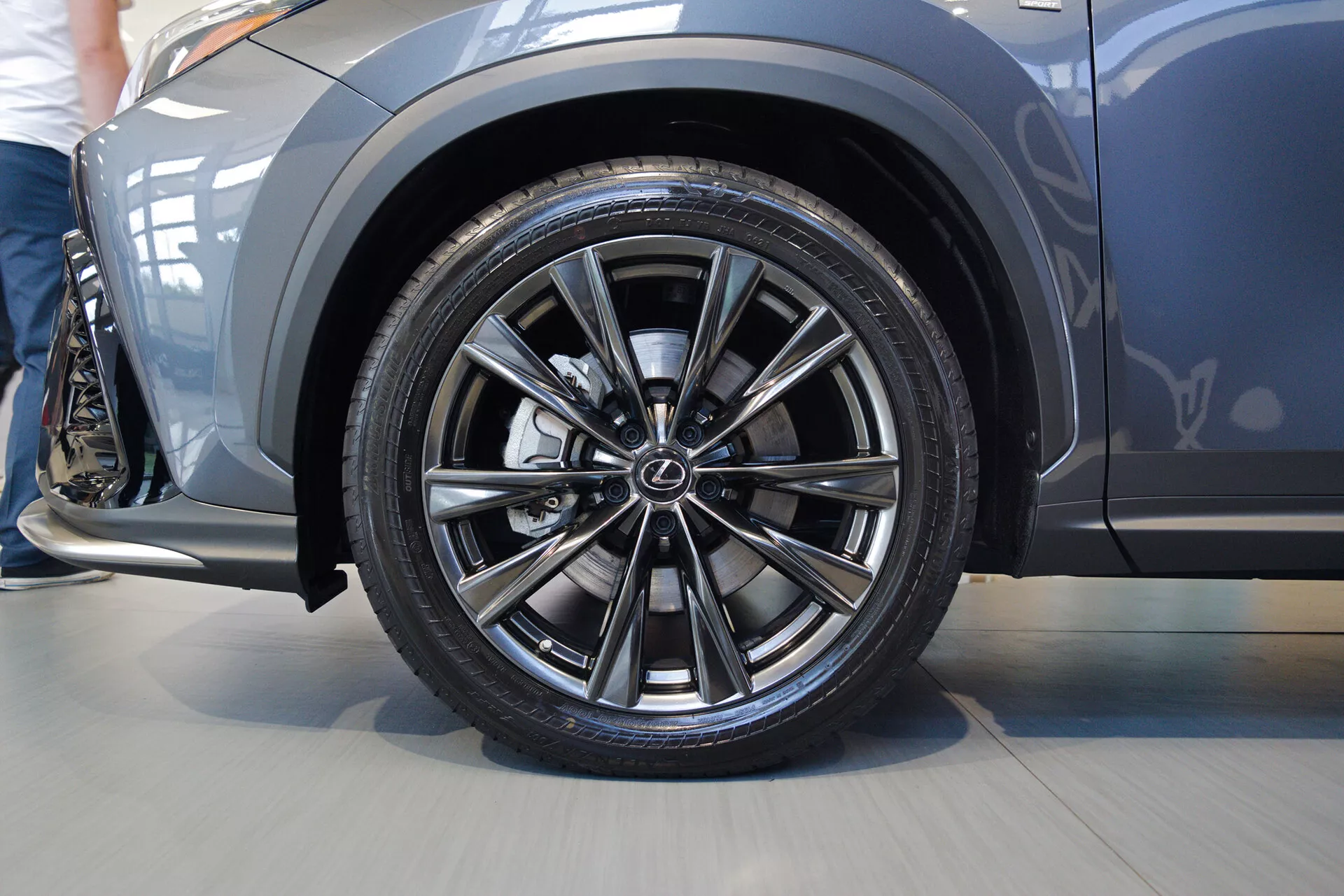 Detailný záber na predné koleso z vodičovej strany Lexus NX 2021, zobrazujúci dizajn zliatinového ráfika s dvojitými lúčmi a logom výrobcu. Nízky profil čiernej pneumatiky zdôrazňuje športový výkon. Nad kolesom je viditeľný mierne vyklenutý, kovový blatník, čo naznačuje športový vzhľad. Bočný panel ukazuje elegantný, aerodynamický dizajn. Obrázok bol nasnímaný v interiéri s blízkym zameraním na koleso a predný bočný panel.