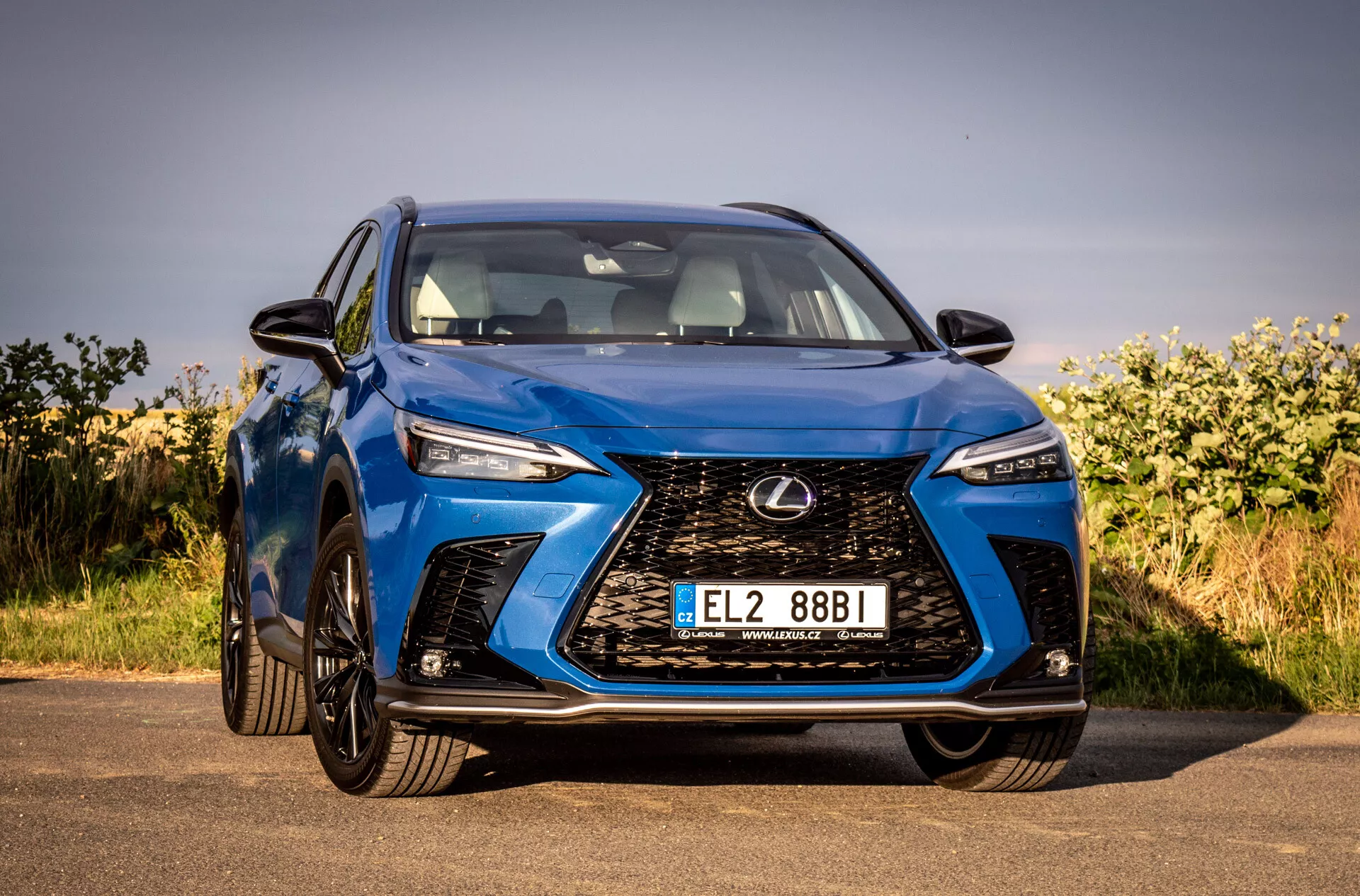 Pohľad z predného uhla na modré SUV Lexus NX 2021 zdôrazňujúci jeho zložitú vretenovú mriežku, uhlové LED svetlomety, lesklý povrch a aerodynamický dizajn, zaparkované vonku pod jasnou oblohou