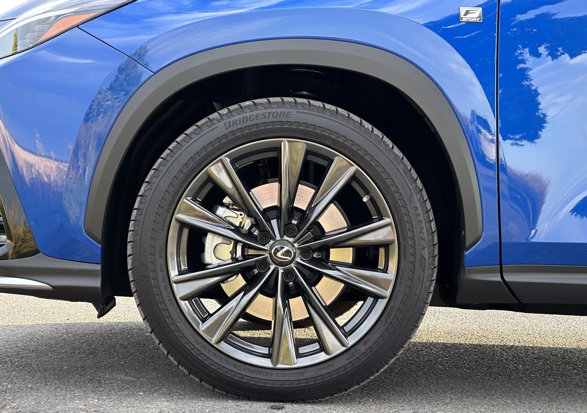 Detailný pohľad na predné koleso na strane spolujazdca Lexus NX 2021 s viacerými drážkami s dvojfarebným povrchom a pneumatikou Bridgestone, na pozadí s živou modrou karosériou