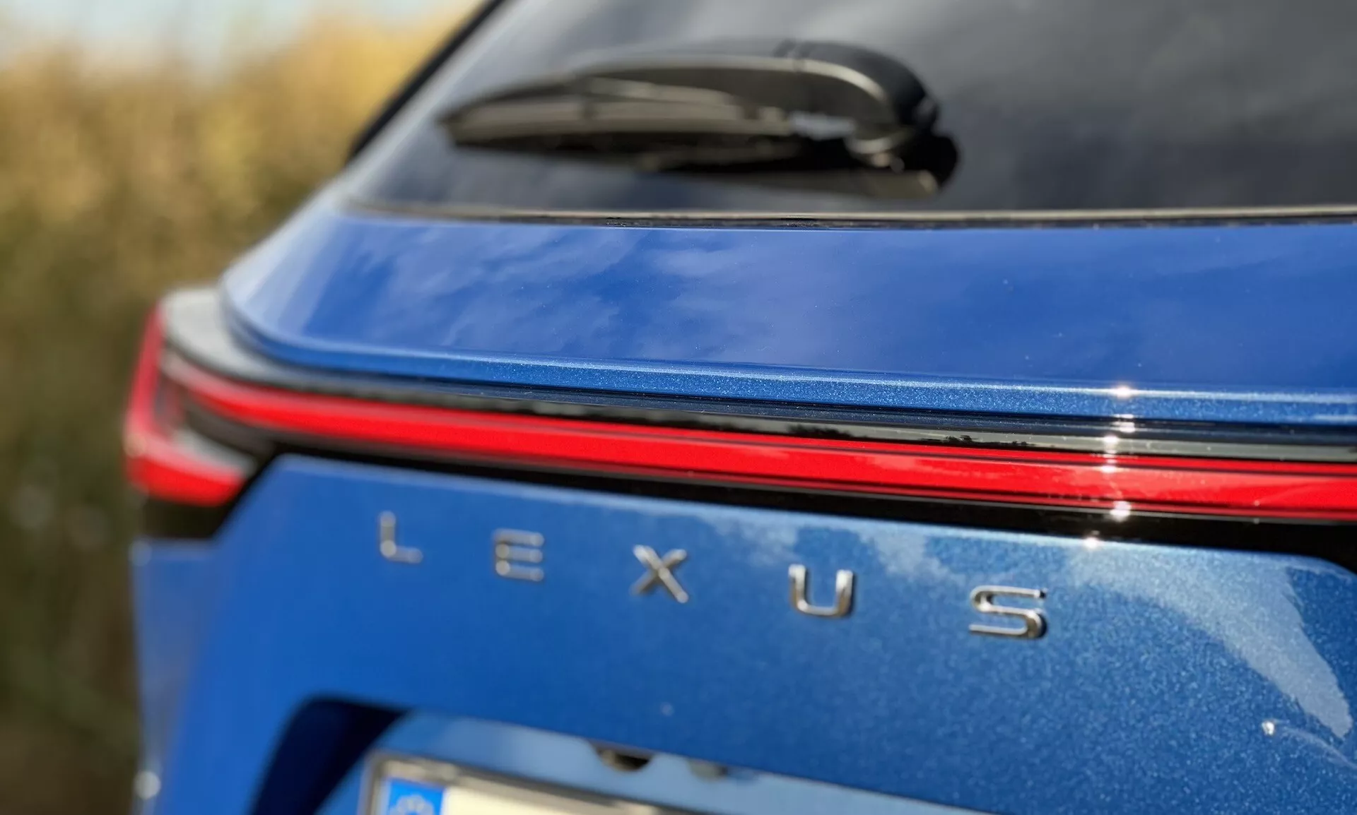 Detailný pohľad na zadnú časť značky vozidla Lexus NX 2021 s strieborným odznakom Lexus, červeným svetelným pásom nad názvom na metalickom modrom telese pri dennom svetle
