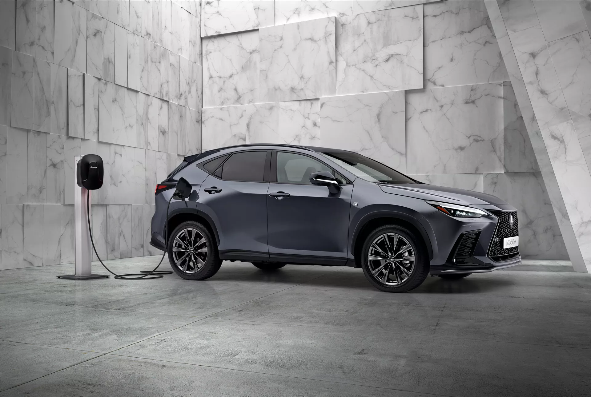 Tento obrázok zobrazuje Lexus NX 2021, moderné kompaktné SUV s elegantným a športovým dizajnom, fotografované z pohľadu trištvrte zepredu. Vidíme ľavú stranu a čiastočne aj prednú časť vozidla, umiestneného diagonálne v minimalistickom prostredí zdobenom veľkými svetlými mramorovými dlaždicami, naznačujúcimi prémiovú atmosféru. Lexus NX sa môže pochváliť výraznými uhlovými LED svetlami a výraznou prednou mriežkou, zdôrazňujúc jeho sofistikovanosť. Viditeľné sú aerodynamicky tvarované bočné zrkadlá, zložité viacramenné zliatinové kolesá a zapustené kľučky dverí, ktoré zdôrazňujú športový a pokročilý dizajn. Dôležitým detailom je pripojenie auta k elektrickej nabíjacej stanici, čo signalizuje, že ide o elektrické vozidlo alebo plug-in hybrid. Nabíjací port na prednom blatníku naznačuje jeho elektrické schopnosti. Tento statický záber sa zameriava na predvedenie dizajnu vozidla a jeho technologických funkcií v pokojnej, vyváženej kompozícii s množstvom negatívneho priestoru, čo stelesňuje exkluzivitu a inováciu v automobilovom dizajne.