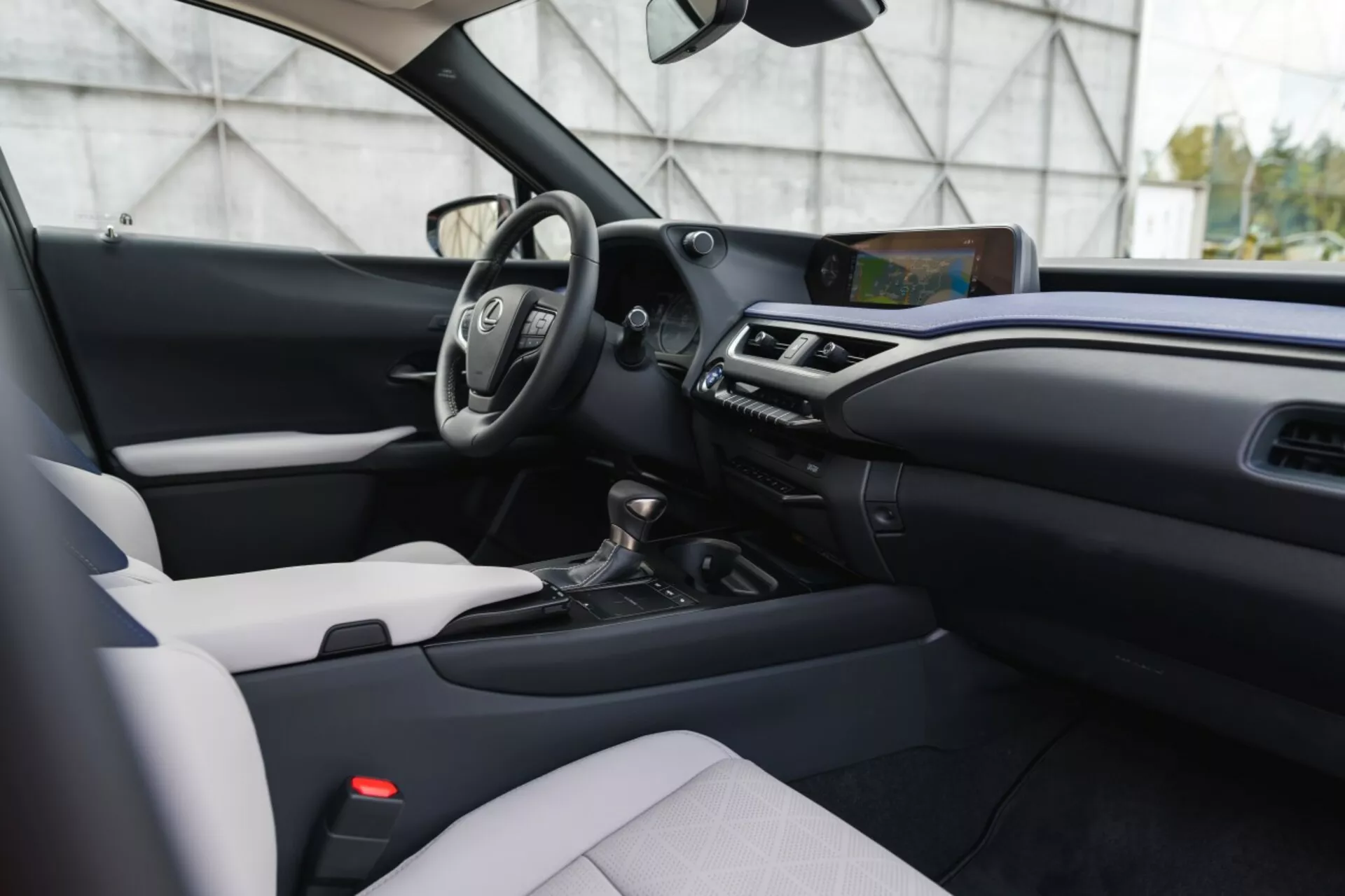 Interiér Lexus UX 2019 z pohľadu predného spolujazdca zobrazujúci palubnú dosku, stredovú konzolu, vodičové sedadlo s volantom a digitálnymi displejmi, dotykový informačno-zábavný systém, klimatizáciu, volič prevodovky, sedadlo spolujazdca čalúnené v dvojfarebnom prevedení a moderné dekoratívne prvky.