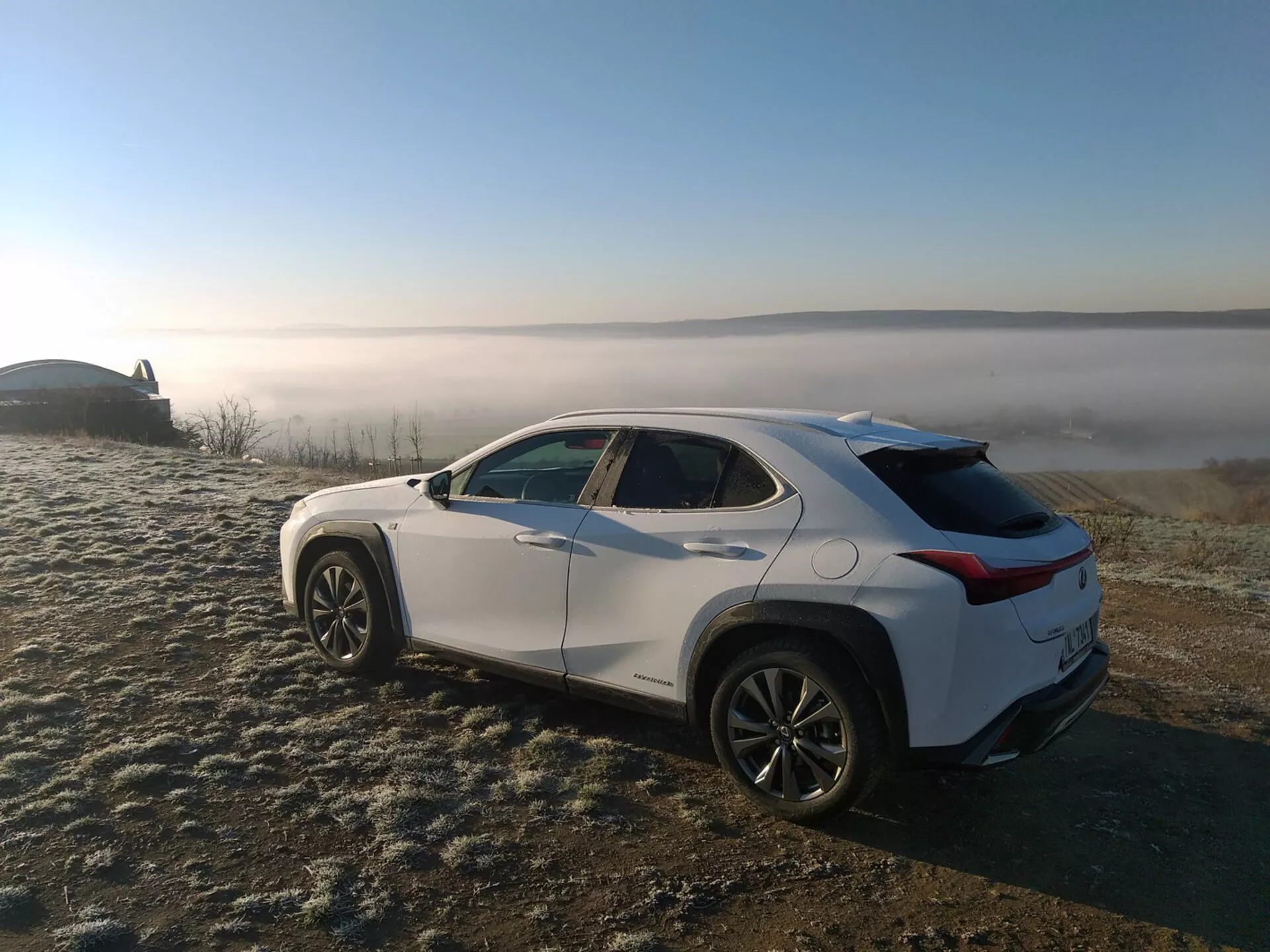Crossover SUV Lexus UX 2019 zaparkovaný na mrazom pokrytej tráve s pozadím hmlistej krajiny, zobrazuje profil vozidla z uhla zozadu tri štvrtiny, čo zdôrazňuje bočné línie karosérie, oblúky kolies, zúženú strechu a zliatinové kolesá.