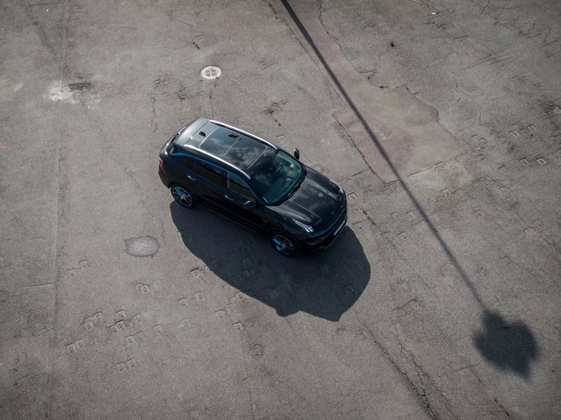 Zvýšený pohľad z vtáčej perspektívy na model Lynk & Co 01 2021, elegantná čierna limuzína s otváracou strechou, umiestnená diagonálne na spevnenej ploche zobrazujúca celú hornú časť, vrátane strechy a kapoty, v ranom rannom alebo neskorom popoludňajšom slnečnom svetle vrhajúcej dlhý tieň
