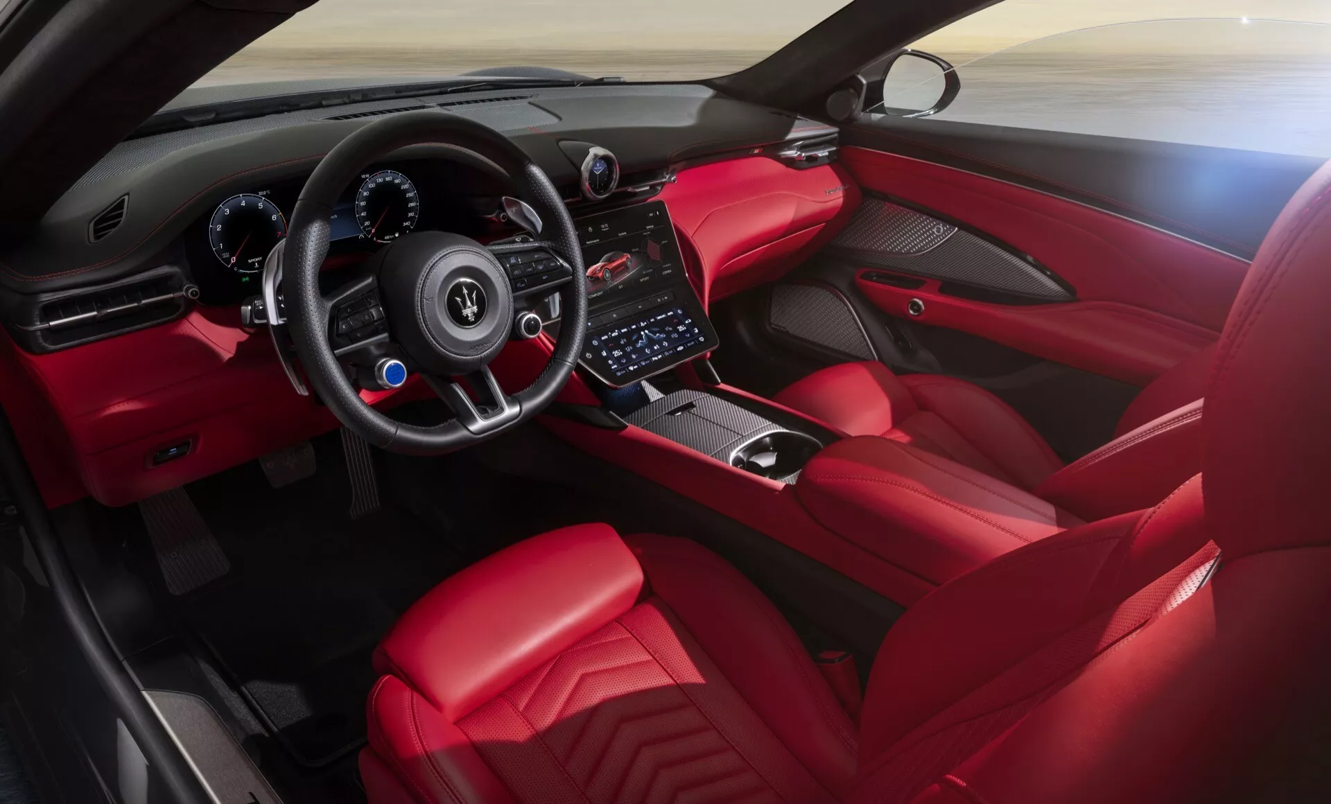 Interiérový pohľad na Maserati GranCabrio 2. generácie roku 2024 zameraný na kokpit a palubnú dosku, obsahujúci športový volant s plochým dnom s tlačidlami ovládania, digitálne prístrojové panely za volantom, dotykovú obrazovku infotainmentu v stredovej konzole, minimalistické tlačidlá klímy, volič prevodovky naznačujúci automatickú alebo manuálnu sekvenčnú prevodovku obklopený ovládacími gombíkmi pre režimy jazdy, a prémiový červený poťah s akcentmi z uhlíkových vlákien a povrchmi z kartáčovaného kovu.