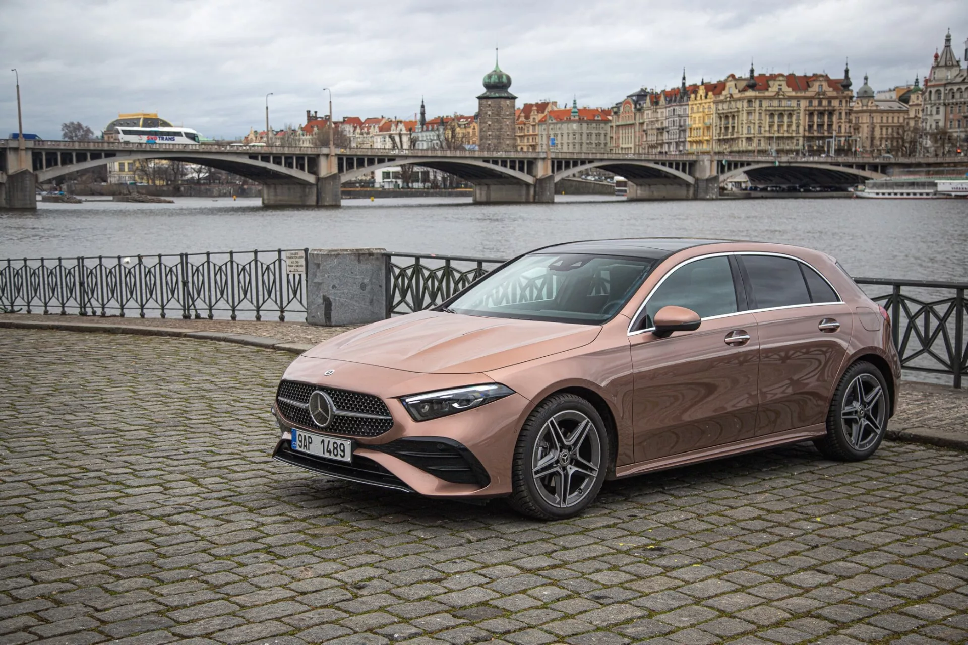 Sedan Mercedes-Benz triedy A z roku 2022 zaparkovaný na európskej dlažobnej ulici s malebným mestským scenériom a vodným pozadím, predvádzajúci jeho štíhly, aerodynamický dizajn, výraznú mriežku, LED svetlomety a metalický náter.