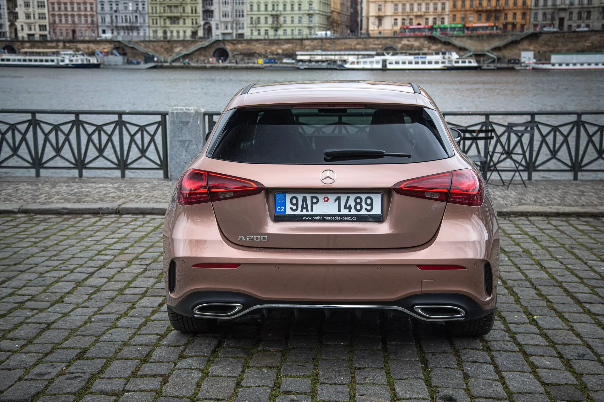 Zadný pohľad na Mercedes-Benz triedy A A 200 zaparkovaný na dlažobnej ploche v mestskom prostredí pri rieke, zobrazujúci dvojitý výfukový systém auta, hatchbackový dizajn a parkovacie senzory