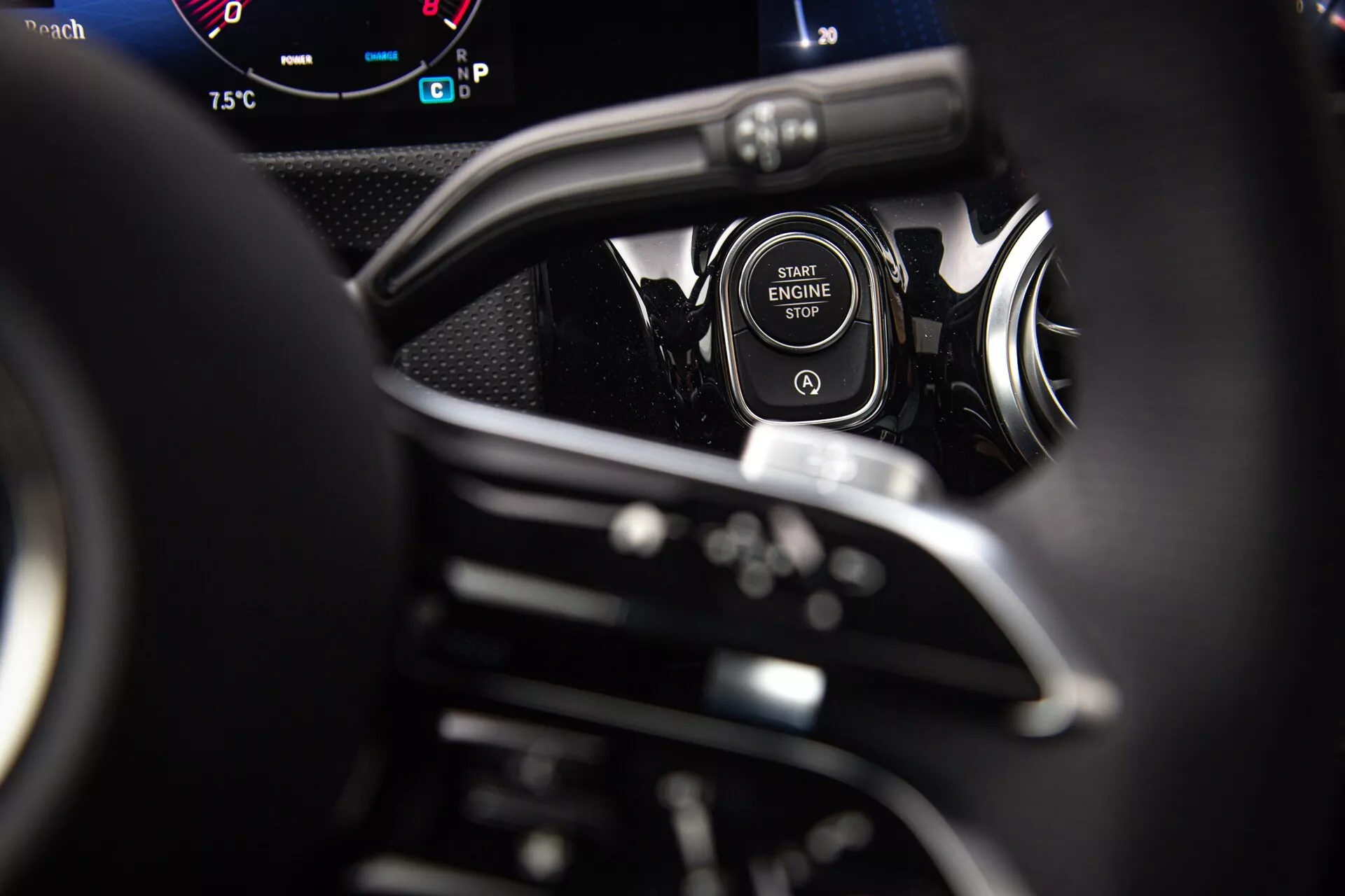 Detailný pohľad na tlačidlo štart/stop motora v Mercedes-Benz triedy A 2022 s chrómovou ozdobnou lištou, čiastočne viditeľným volantom, pádlami na radenie a digitálnymi ukazovateľmi palubnej dosky z pohľadu vodiča
