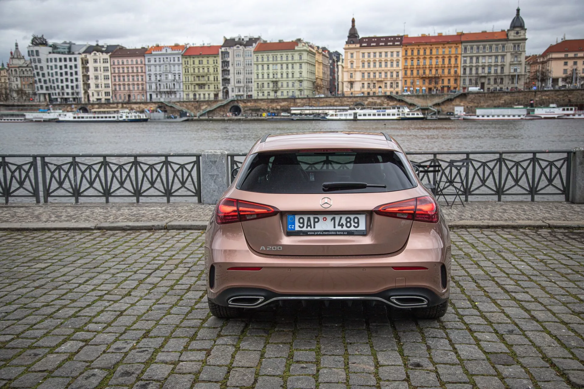 Zadný pohľad na hatchback Mercedes-Benz triedy A z roku 2022 zaparkovaný na dlažobnej ploche s vodou a budovami na pozadí pod zamračenou oblohou, s LED zadnými svetlami, spojlerom na streche a dvojitými výfukovými rúrami