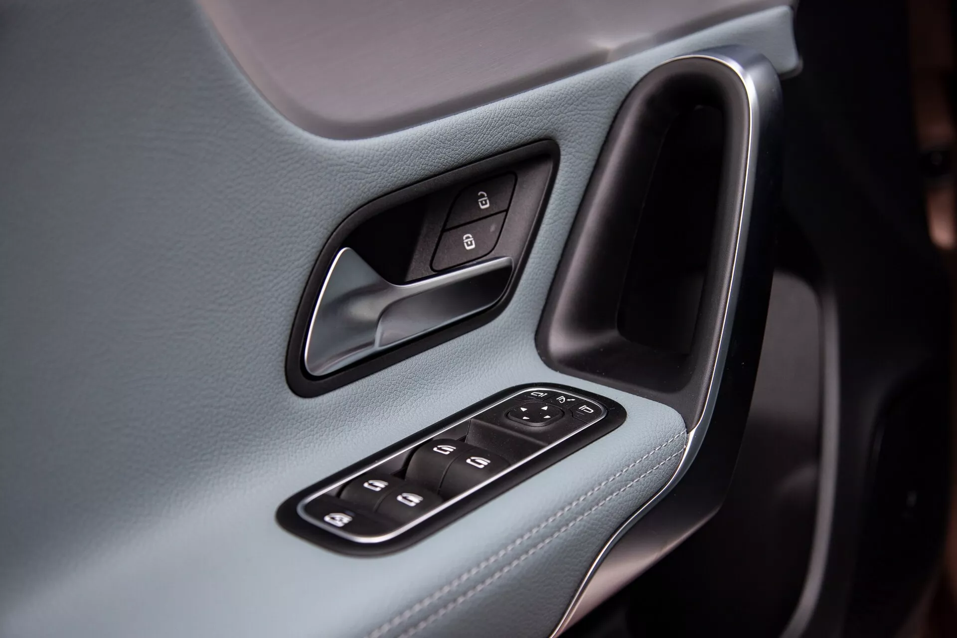 Detailný interiérový záber ovládacieho panela dverí vodiča Mercedes-Benz triedy A 2022, zdôrazňujúci ergonomickej design a technologické funkcie ako ovládanie zrkadiel a okien, chromovaná kľučka dverí proti koženému poťahu.