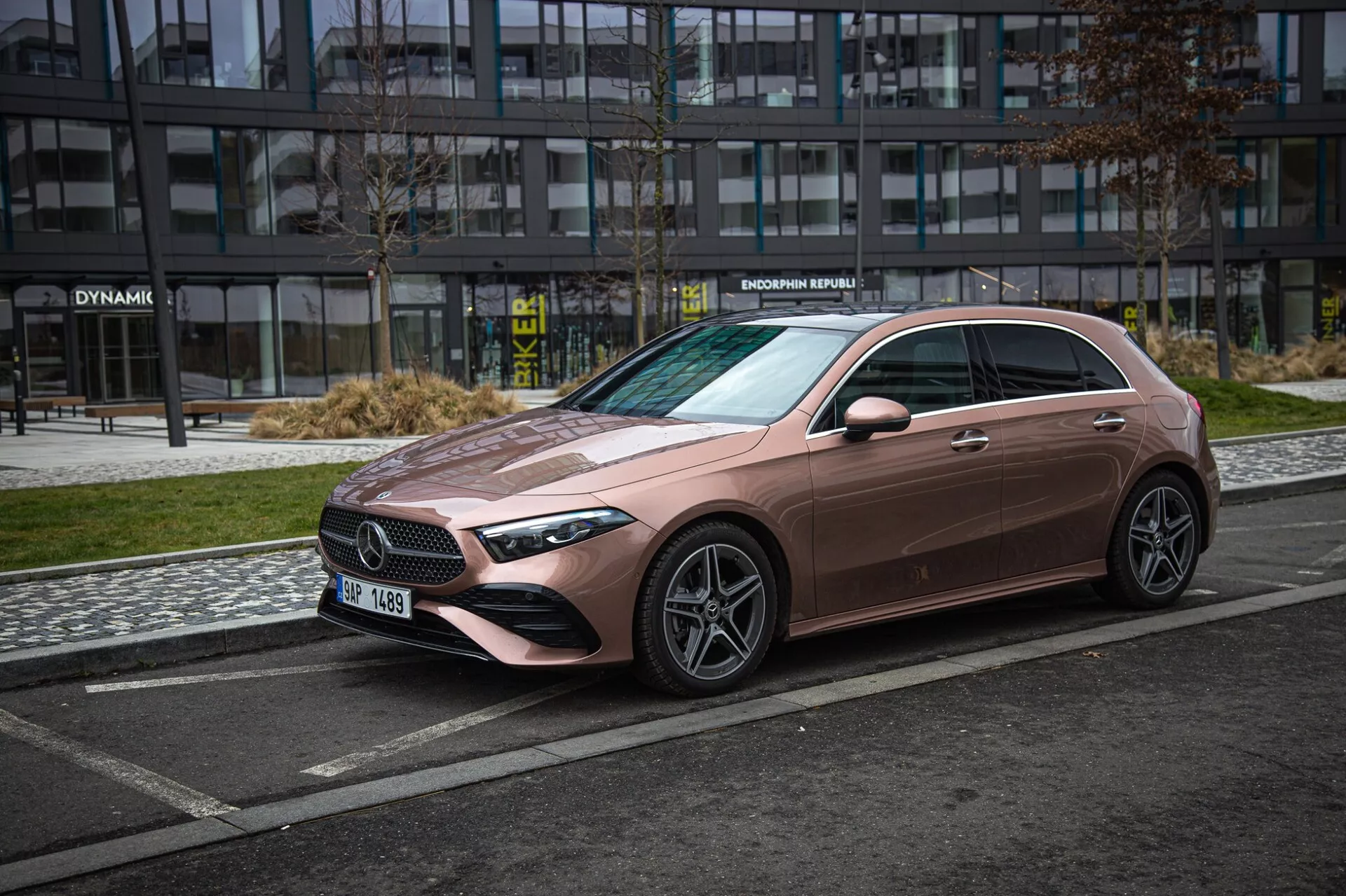 Sedan Mercedes-Benz triedy A z roku 2022 v jedinečnom metalickom ružovom odtieni, zobrazený z uhla troch štvrtín spredu, ktorý zdôrazňuje jeho elegantnú prednú mriežku, LED svetlomety, tvarovanú kapotu a kolesá zo zliatiny s viacero lúčmi na mestskom pozadí