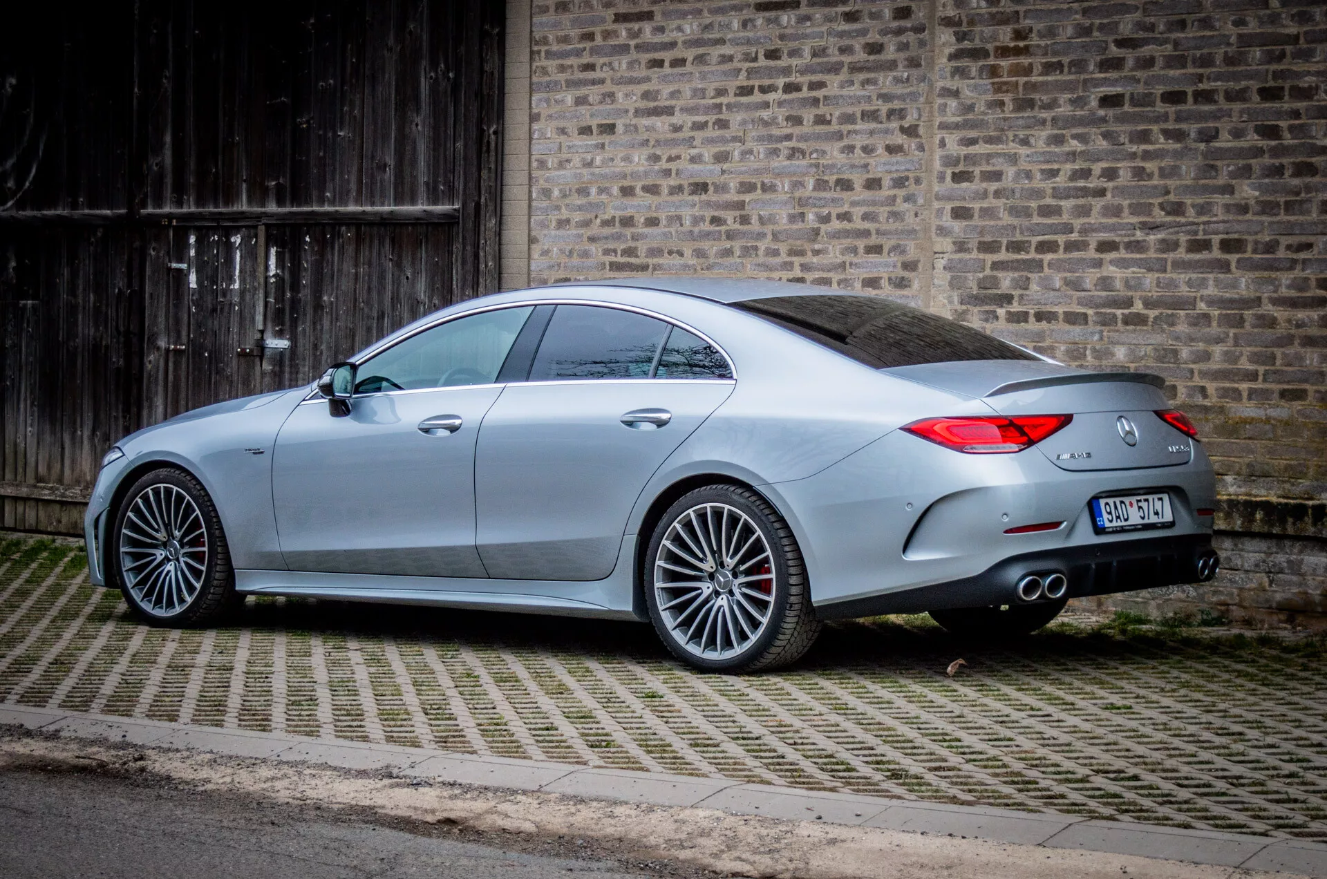 Mercedes-Benz CLS z roku 2021 (CLS-AMG Coupe) zaparkované pod uhlom na dlažobnom príjazde vedľa budovy s drevenou bránou, ukazuje jeho zadný trojštvrťový pohľad s viditeľnými dizajnovými čiarami, LED zadnými svetlami, dvojitým výfukom, veľkými zliatinovými kolesami s viacerými spicami a pokročilými technologickými funkciemi ako senzory parkovania vzadu a integrované smerové signály na bočných zrkadlách, v mestskom alebo polomestskom prostredí.