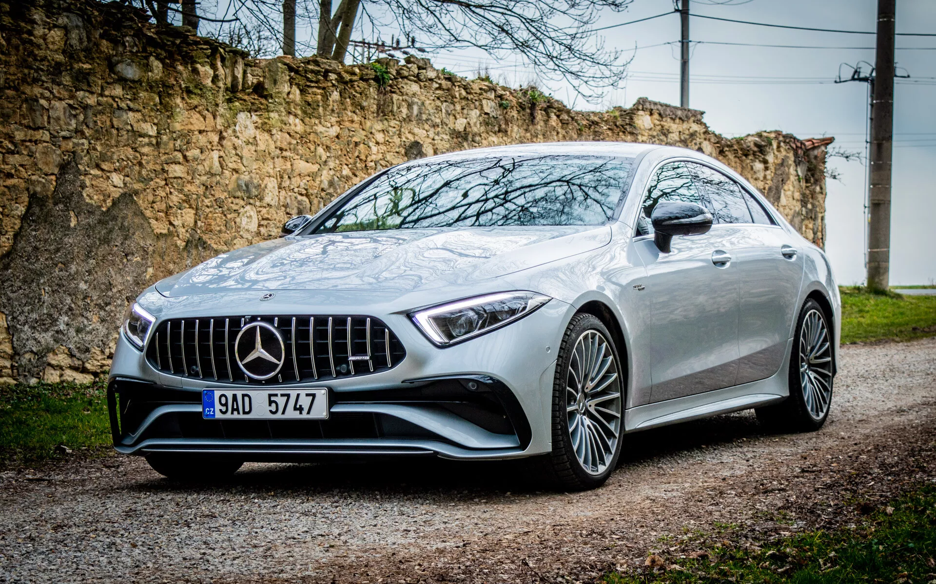 Strieborné kupé Mercedes-Benz CLS AMG 2021 z trojštvrtečného predného uhla ukazujúce mriežku, LED svetlomety, predný nárazník a kolesá z ľahkých zliatin na štrkovej ceste s rustikálnym pozadím