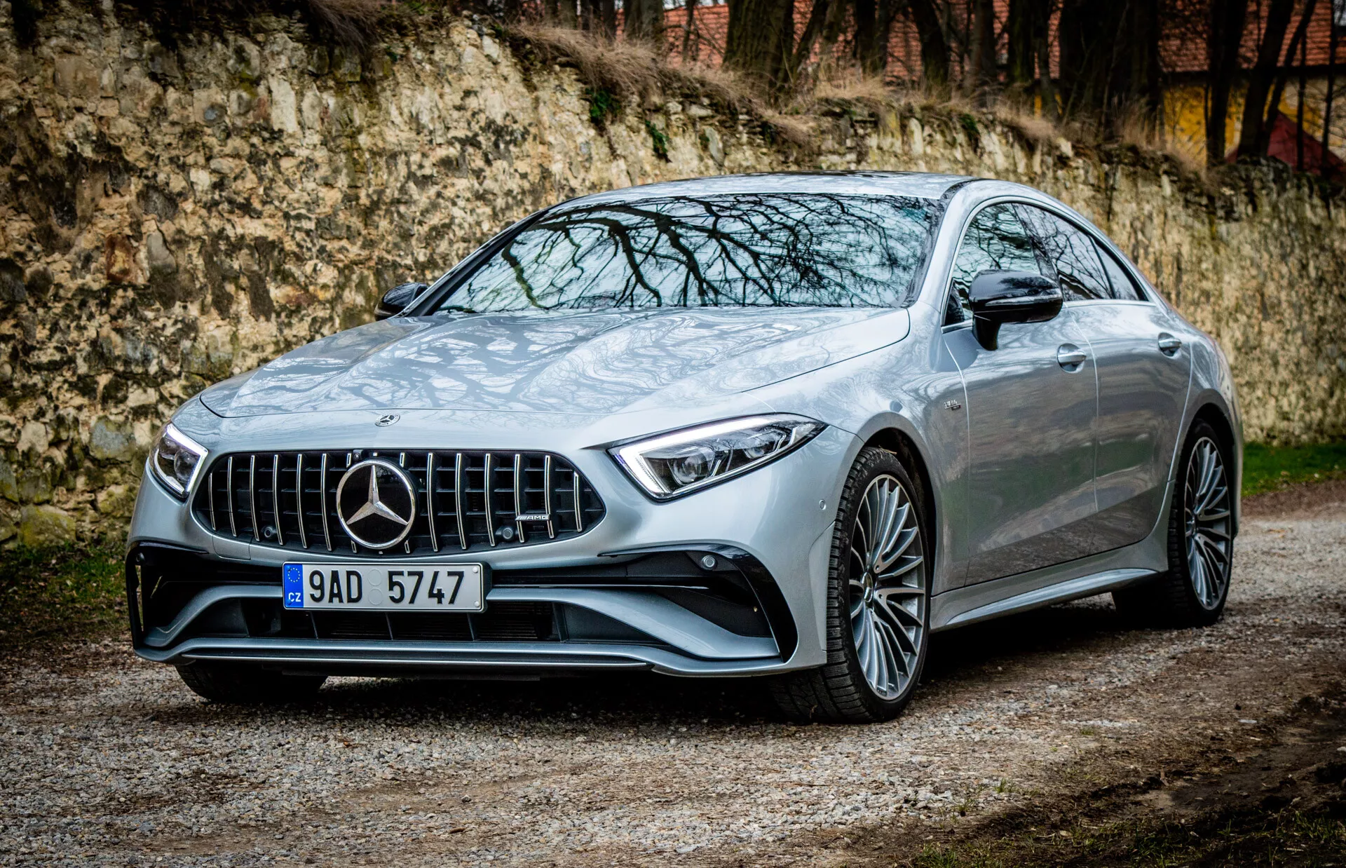 Mercedes-Benz CLS-AMG Coupe 2021 zaparkované na štrku s vegetáciou a architektonickou štruktúrou na pozadí, predvádzajúce jeho charakteristickú mriežku, LED predné svetlomety, aerodynamický nárazník, športové línie kapoty, viaceré zliatinové kolesá, zapustené kľučky dverí a potenciálny panoramatický strešný okno v luxusnom kovovom prevedení