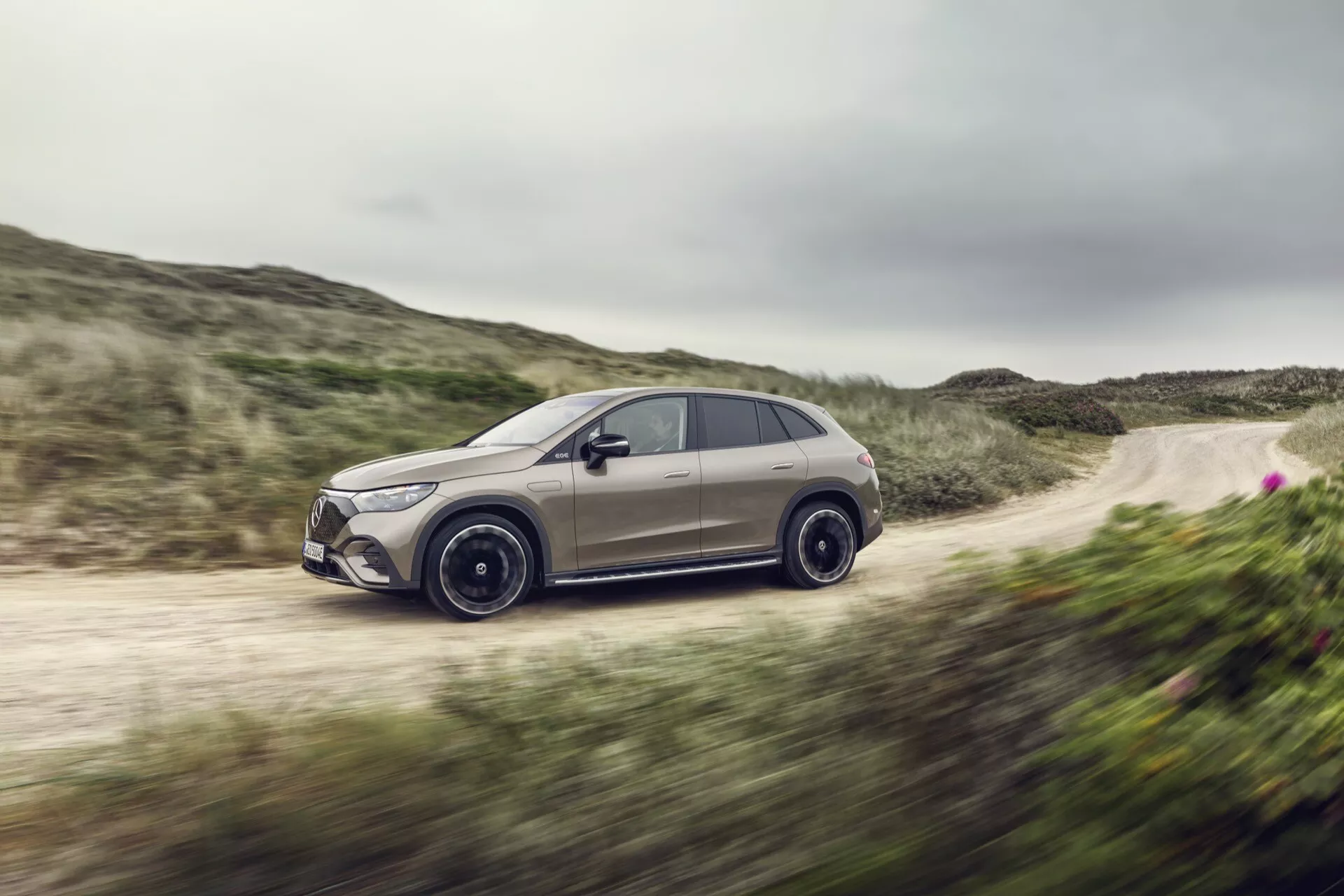Mercedes-Benz EQE-SUV 2023 v dynamickom pohybe na neupravenej ceste, predvádzajúci aerodynamický dizajn, športové zliatinové kolesá, bezpečnostné bočné zrkadlá so zabudovanými smerovkami a prémiové tónované okná, ilustrujúce pokročilé technologické funkcie a schopnosť zvládať rôzne terény.