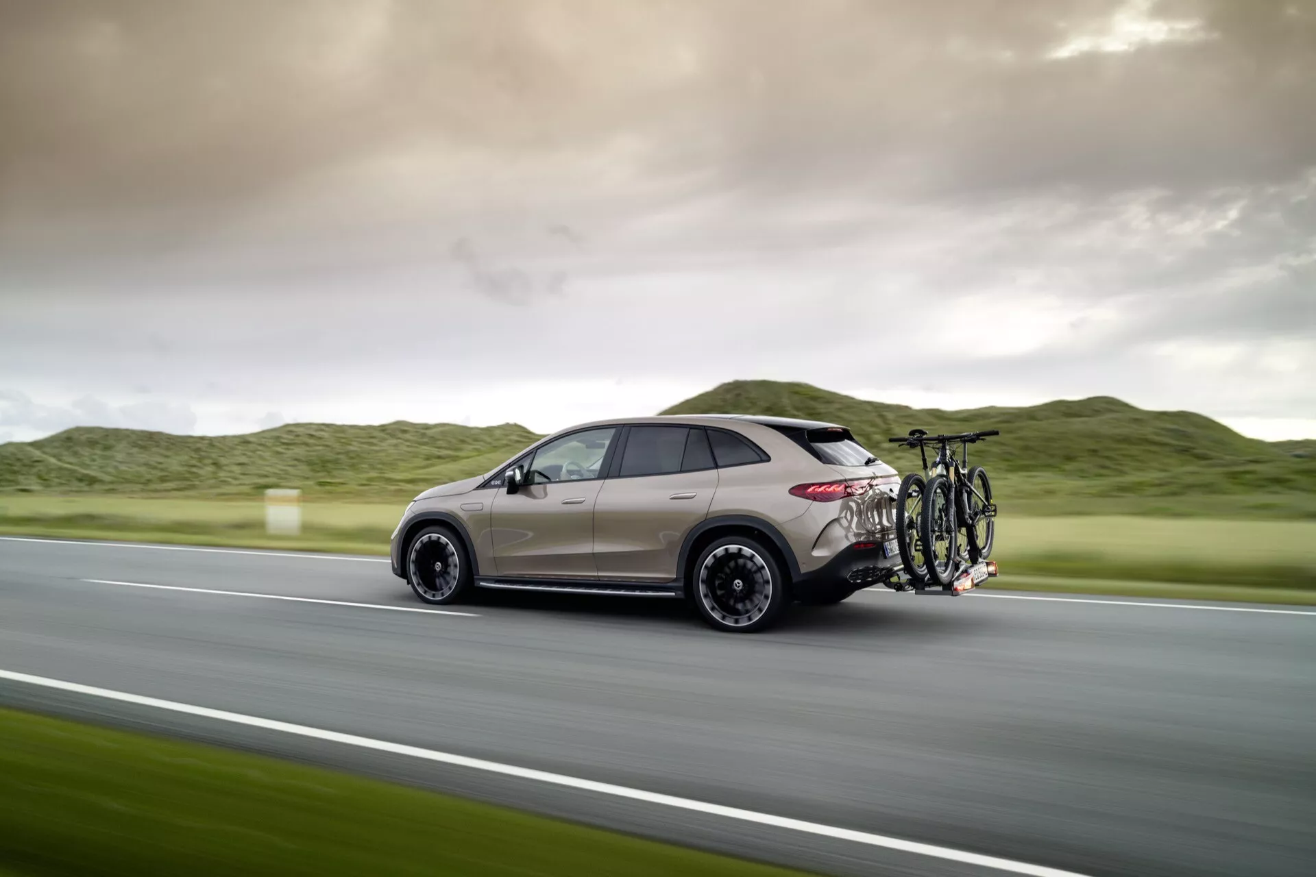 SUV Mercedes-Benz EQE 2023 v pohybe, bočný zadný pohľad zvýrazňujúci aerodynamický dizajn, metalickú béžovú farbu a vybavenie nosičom bicyklov s dvoma bicyklami