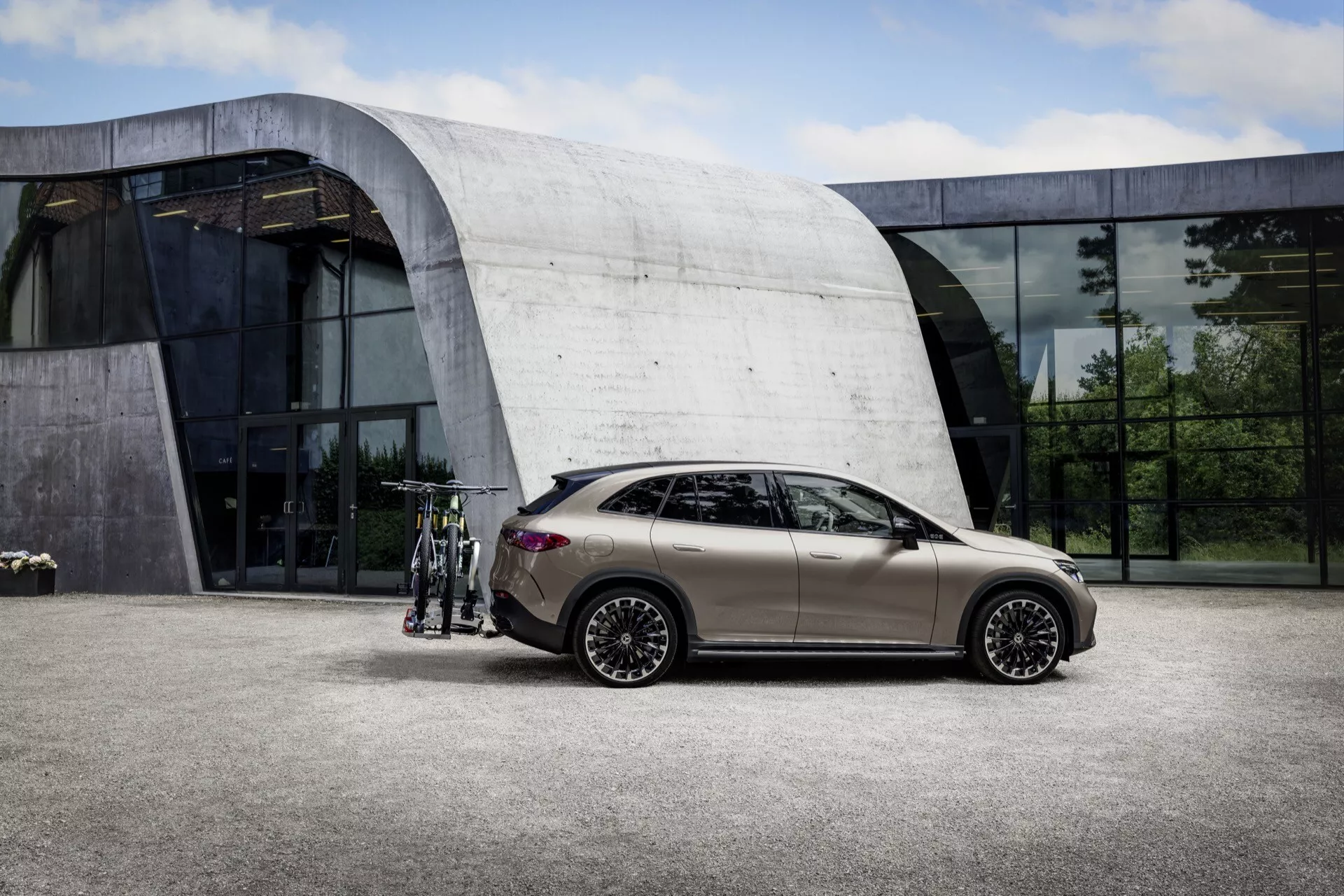 Model SUV Mercedes-Benz EQE 2023 v matnej bronzovej farbe, predstavujúci moderný hatchback dizajn a zložité zliatinové kolesá z bočného profilu, naznačujúci pokročilé technologické vlastnosti ako bezkľúčový vstup, zaparkovaný vedľa súčasnej budovy so sklenenými panelmi.