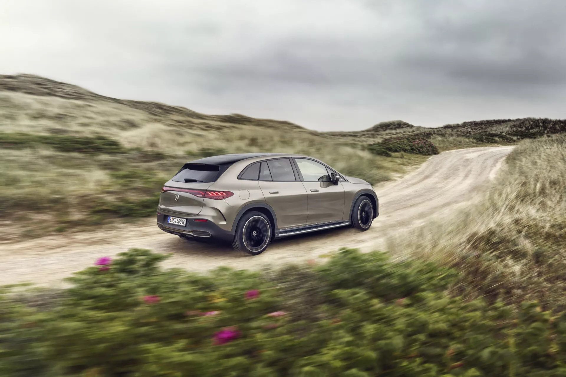 Mercedes-Benz EQE-SUV 2023 v pohybe na nezpevnenej ceste, s pohľadom z zadného štvrťúhla, zliatinovými kolesami, LED zadnými svetlami a výkonnostným dizajnom na pozadí prírodného krajiny.