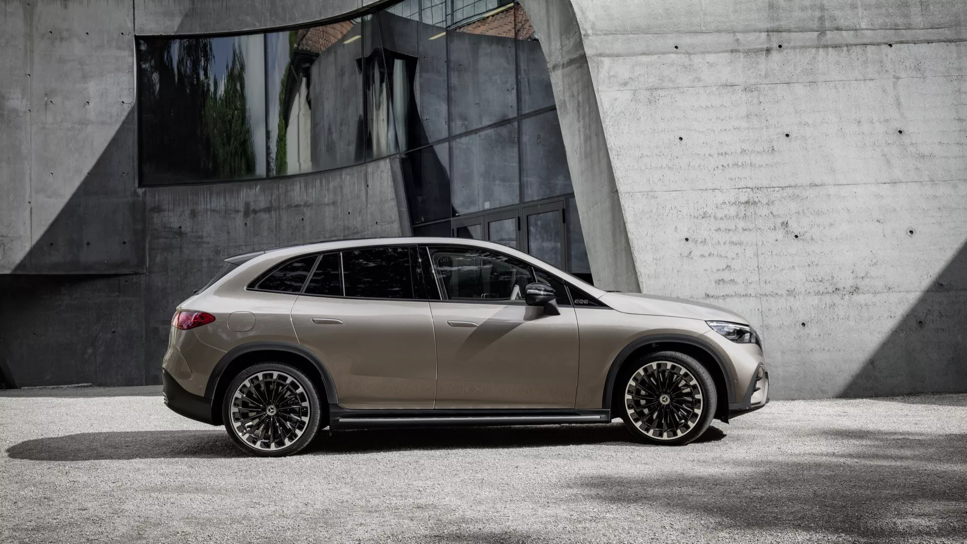 Celý bočný profil moderného hatchbacku Mercedes-Benz EQE-SUV 2023 v matnej béžovej farbe, s detailne spracovanými zliatinovými kolesami s dizajnom špic, kľučkami dverí v farbe karosérie, integrovanými smerovými signálmi na bočných zrkadlách a čiernym lemovaním okien, zaparkovaný na betóne v mestskom prostredí