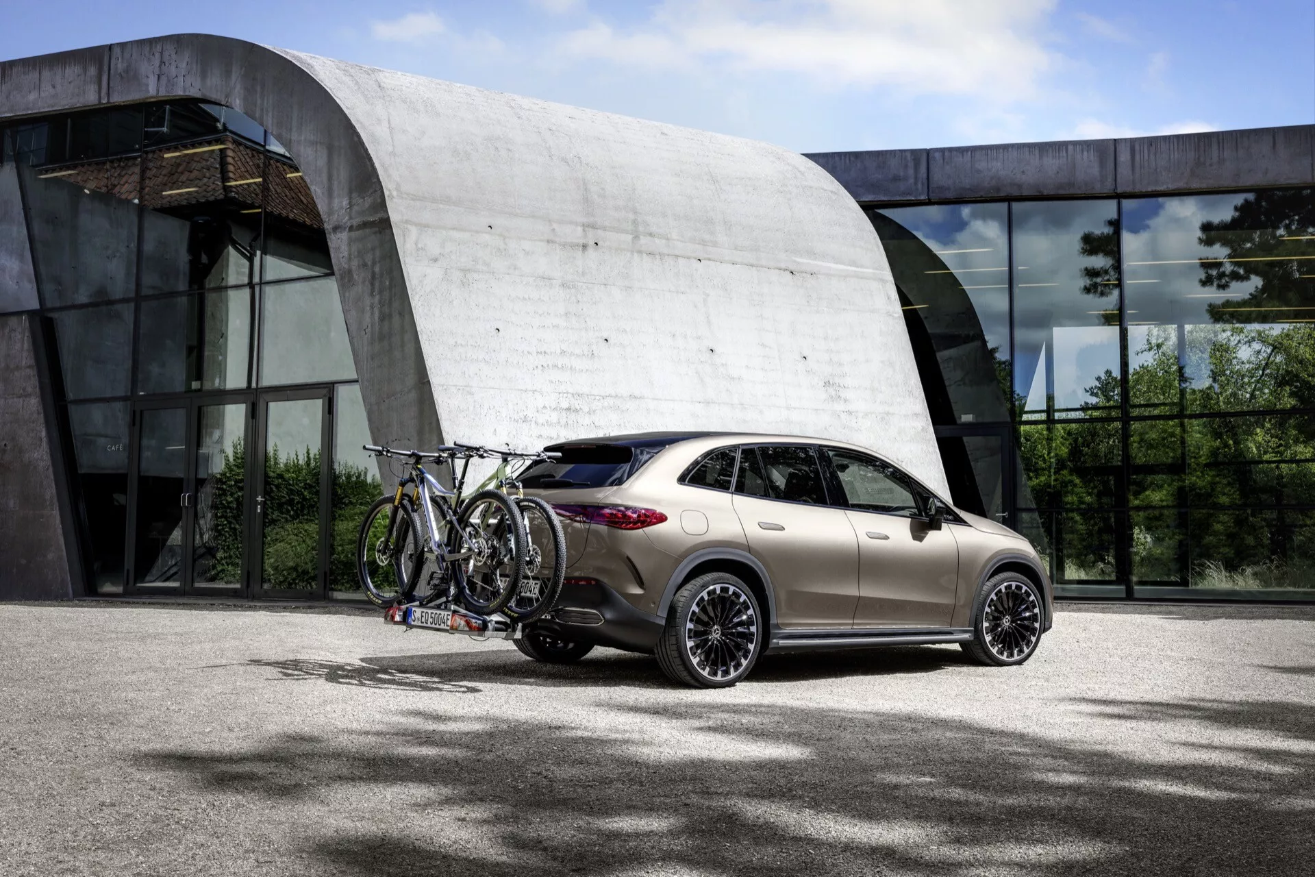 Mercedes-Benz EQE-SUV 2023 vo svetlozlatej farbe zaparkovaný vonku s elegantným dizajnom hatchback, moderným LED osvetlením, aerodynamickými bočnými zrkadlami, zliatinovými kolesami s viacerými lúčmi s nosičom bicyklov na streche v jasnom dennom svetle s modernou budovou na pozadí
