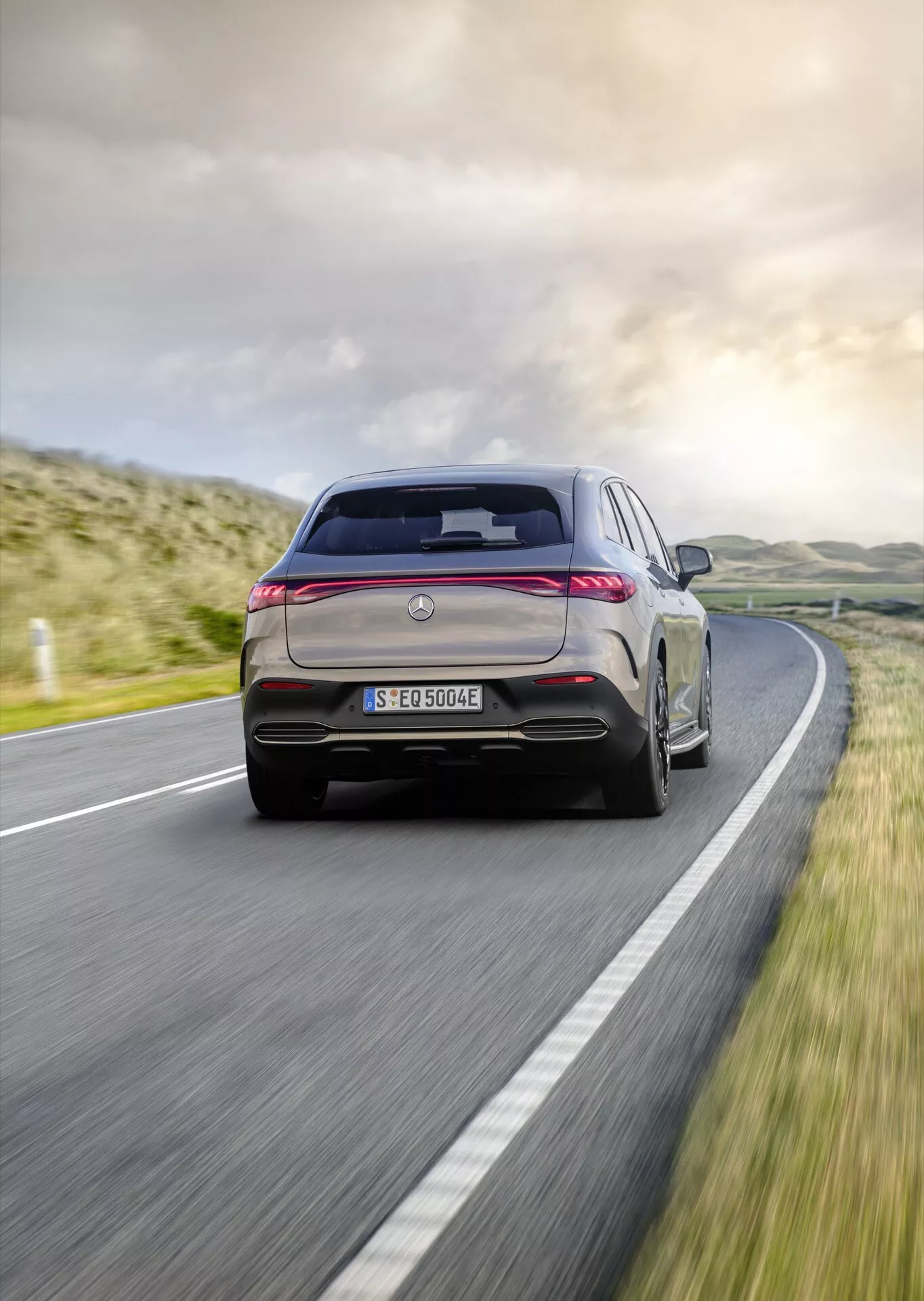 Mercedes-Benz EQE-SUV 2023 v pohybe na vidieckej ceste predvádzajúce zadné LED svetlá, dvojité výfuky, aerodynamický difúzor, integrovaný spoiler, anténu v tvare plutvy žraloka, emblém pre elektrický alebo hybridný model, zadný stierač a prémiové zliatinové kolesá s viacerými ramenami