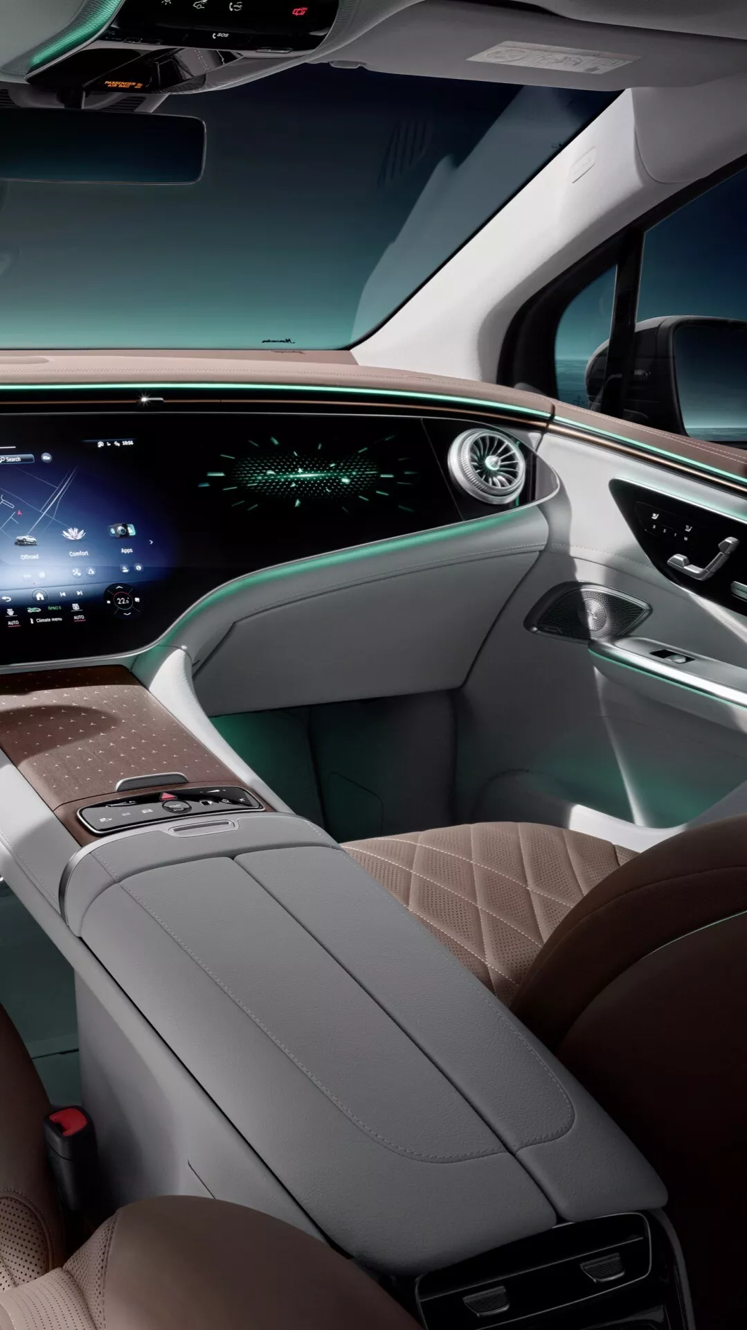 Interiér Mercedes-Benz EQE-SUV 2023 predstavujúci luxusný pohľad na prednú kabinu z pohľadu zadného sedadla, zdôrazňujúci sedadlá z vysoko kvalitnej svetlej kože s diamantovým stehom, elegantná stredová konzola s dreveným povrchom a minimom tlačidiel, rozsiahle horizontálne displeje pre informácie vodiča a infotainment, moderný volant s tlačidlami ovládania a ambientné osvetlenie zdôrazňujúce vysokotechnologický a ergonomický dizajn