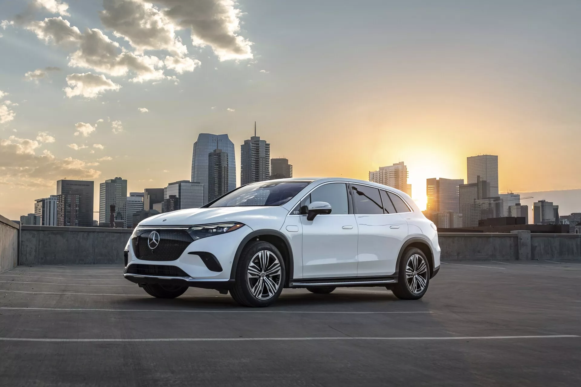 Elegantný biely Mercedes EQS-SUV 2022 zaparkovaný s panorámou mesta a zapadajúcim slnkom v pozadí. Uhol predného trojštvrtinového pohľadu ukazuje charakteristickú výraznú mriežku s detailným sieťovým vzorom a centrálnym emblémom, elegantné LED svetlomety, dvojité sacie otvory alebo vetracie otvory a veľké viacramenné zliatinové kolesá. Jemne sklonená strecha a zvýšená výška svetlosti naznačujú dizajn SUV. Bočné zrkadlá pravdepodobne obsahujú technologické funkcie, ako sú integrované smerové svetlá alebo kamery na sledovanie mŕtveho uhla. Obrázok je nádherne osvetlený jemnými odleskami zapadajúceho slnka, ktoré zvýrazňujú dizajnové línie vozidla. Mestské prostredie s mrakodrapmi dodáva moderný a sofistikovaný nádych, čím naznačuje cieľovú skupinu mestských spotrebiteľov, ktorí oceňujú štýl a pokročilé technológie.