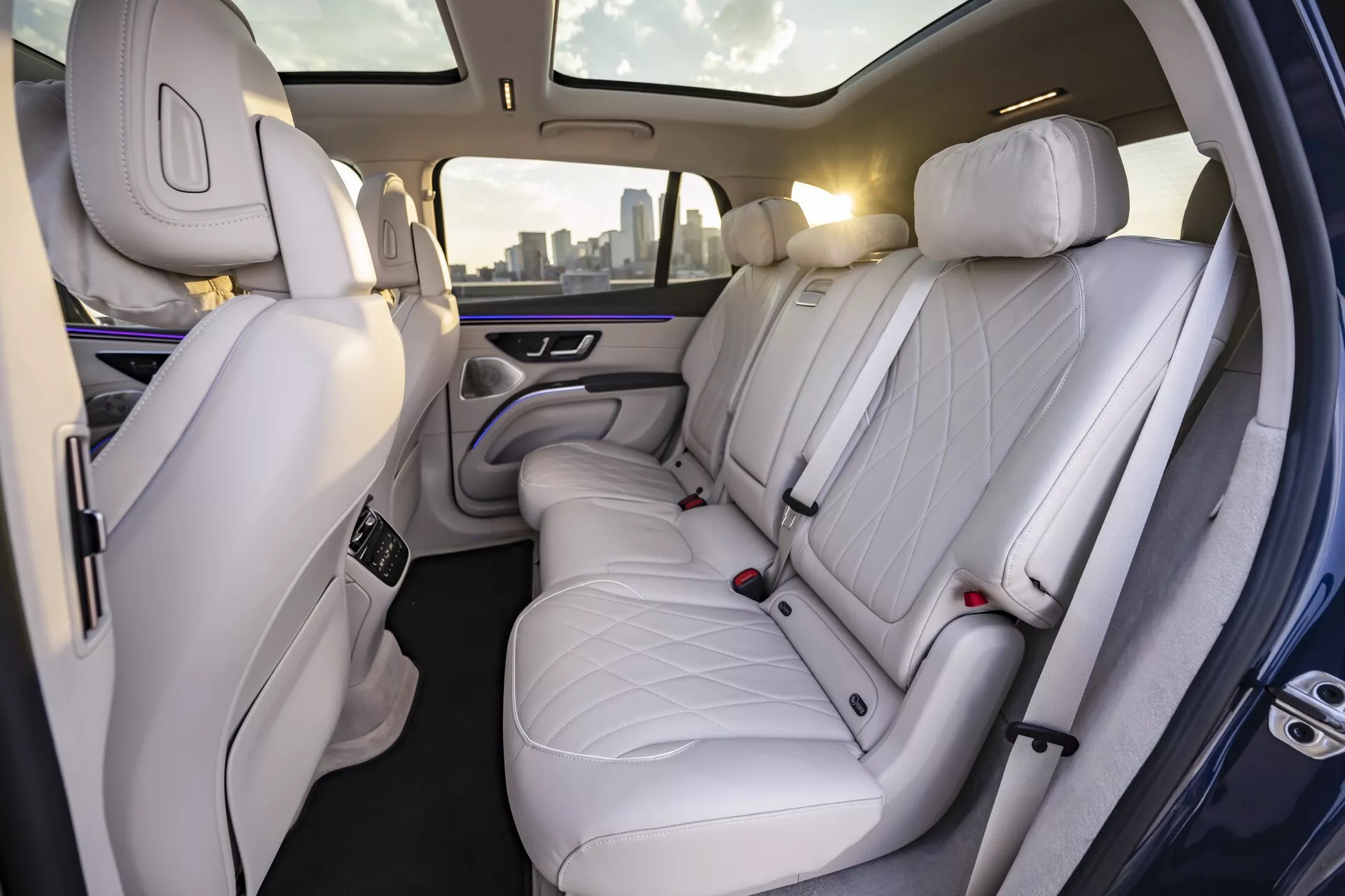 Pohľad na interiér Mercedes EQS-SUV 2022, ktorý zobrazuje luxusný zadný priestor pre cestujúcich. Obrázok, urobený z uhla mierne nakloneného doprava, odhaľuje vysoko kvalitné biele alebo svetlosivé kožené sedadlá so stehmi v tvare diamantov. S tromi opierkami hlavy poskytujú sedadlá dostatok miesta, čo naznačuje dôraz na pohodlie a eleganciu. Vzadu je viditeľná aj časť sedadla spolujazdca, tiež potiahnutého prémiovou kožou. Sofistikované ambientné osvetlenie s jemným fialovým žiarením pozdĺž dverových panelov a stredovej opierky zlepšuje pocit prémiovosti. Stredová opierka obsahuje ovládacie prvky pravdepodobne na nastavenie klímy a sedadiel a dva držiaky na nápoje.