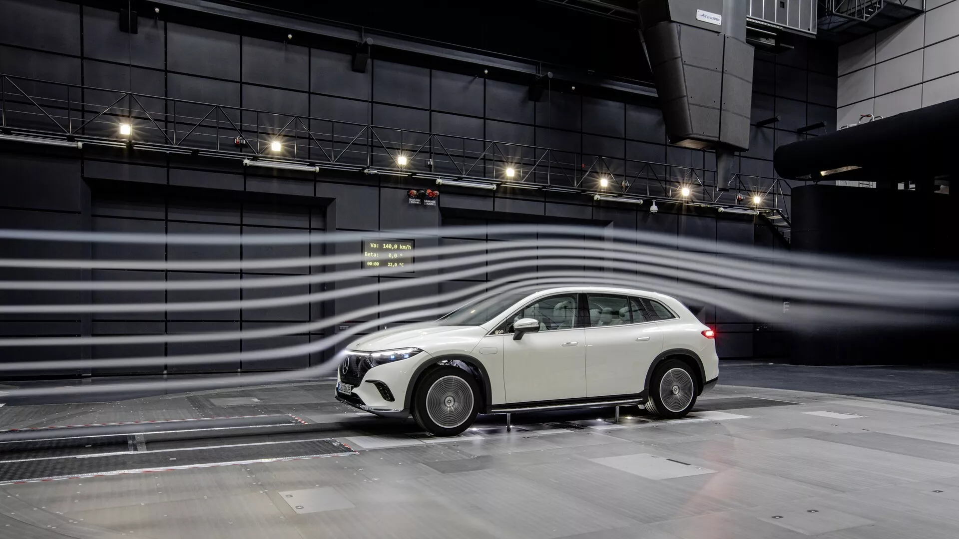 Obraz zobrazuje bočný profil Mercedes EQS-SUV 2022, zdôrazňujúc jeho moderný dizajn v kontrolovanom testovacom zariadení. Vozidlo hatchback alebo kombi model má výraznú prednú mriežku, elegantné bočné zrkadlá, viacpaprskové zliatinové kolesá, integrované hmlové svetlá v prednom nárazníku a strechou namontovanú anténu. Aerodynamické línie a vizualizované prúdenie vzduchu okolo vozidla naznačujú zameranie na efektívnosť paliva a výkon, pričom mokrý povrch odrážajúci vozidlo môže naznačovať testovanie interakcie s povrchom cesty. Usporiadanie naznačuje pokročilé technológie a aerodynamické testy, pravdepodobne pre funkcie ako aktívna aerodynamika, asistenčné systémy pre vodiča a kontrola trakcie.