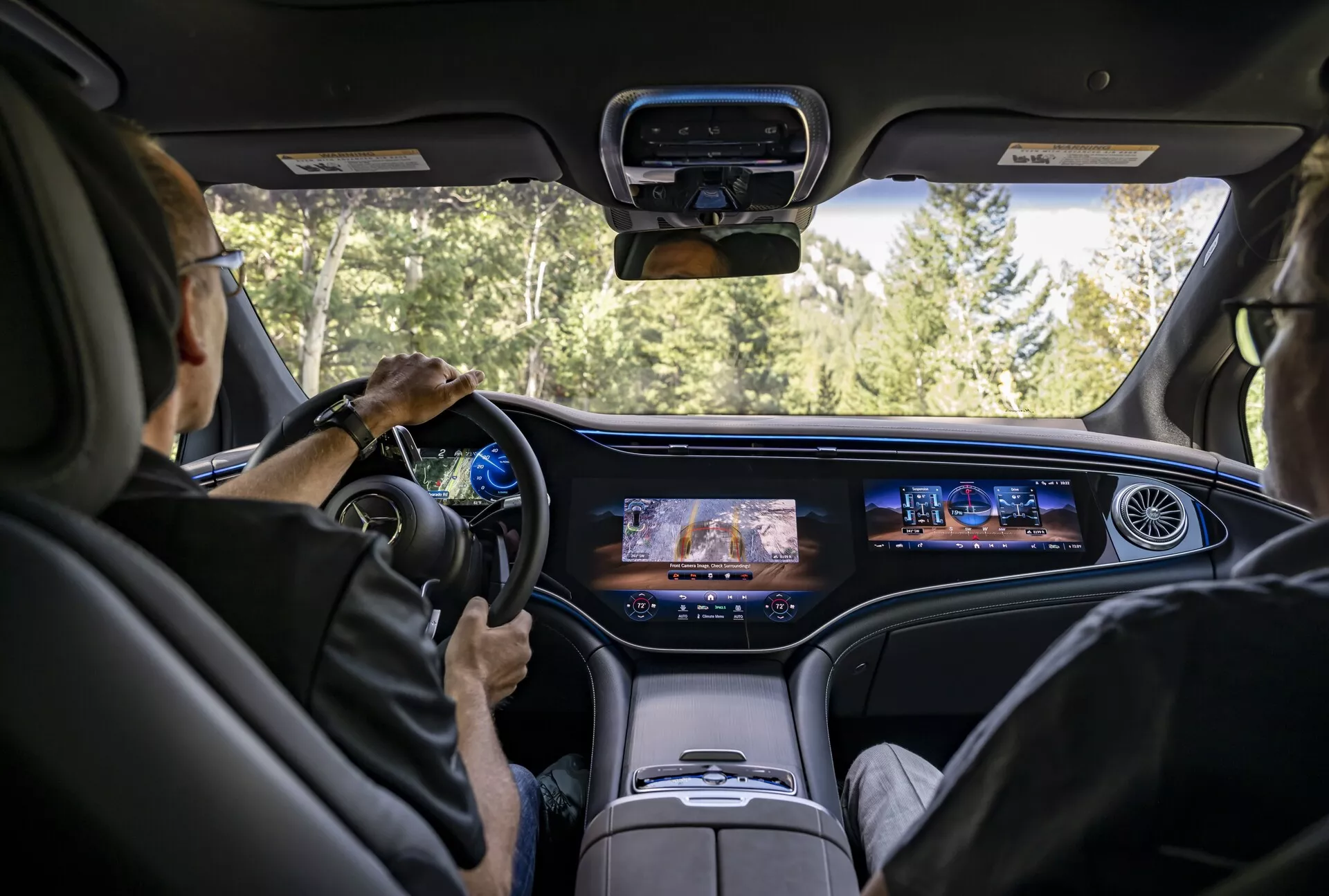 Pohľad do interiéru Mercedes EQS-SUV 2022 zo stredu zadného sedadla, ktorý zobrazuje modernú palubnú dosku s neprerušeným digitálnym displejom, súčasný volant s montovanými ovládacími prvkami, luxusné svetlé kožené sedadlá a zvýšenú stredovú konzolu. Čelné sklo ponúka výhľad na cestu obklopenú lesom, čo naznačuje, že auto je v pohybe.