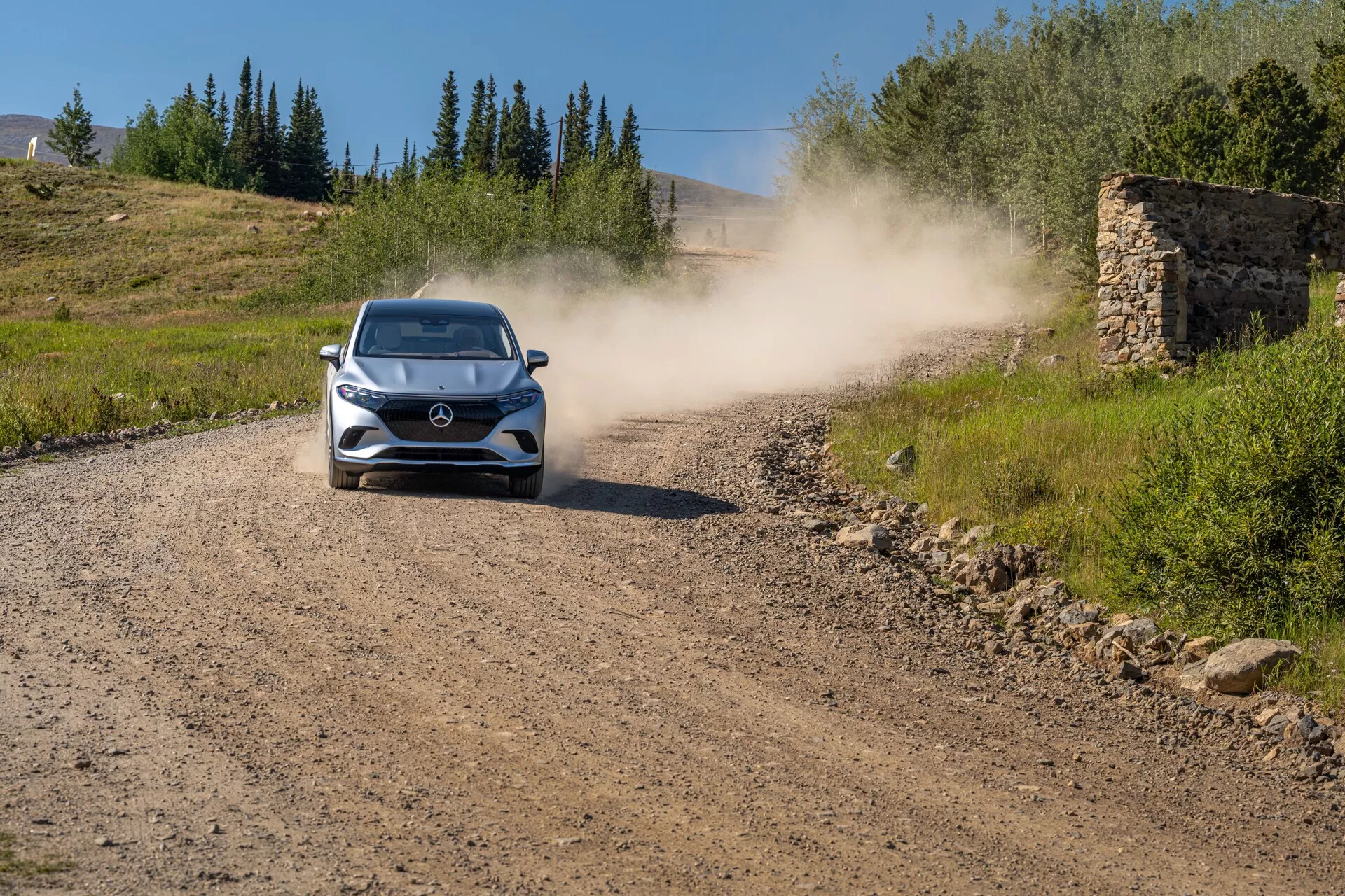 Mercedes EQS SUV 2022 – Moderný hatchback alebo kompaktné SUV, ktoré ide smerom ku kamere na štrkovej ceste, za sebou zanechávajúc prachový oblak. Auto je umiestnené v strede a výrazne ukazuje prednú mriežku, svetlomety, kapotu, čelné sklo a časť strechy. Prostredie naznačuje vidiecku alebo terénnu oblasť s trávou po oboch stranách, jasnou oblohou a zvyškami staršej kamennej štruktúry v pozadí.
