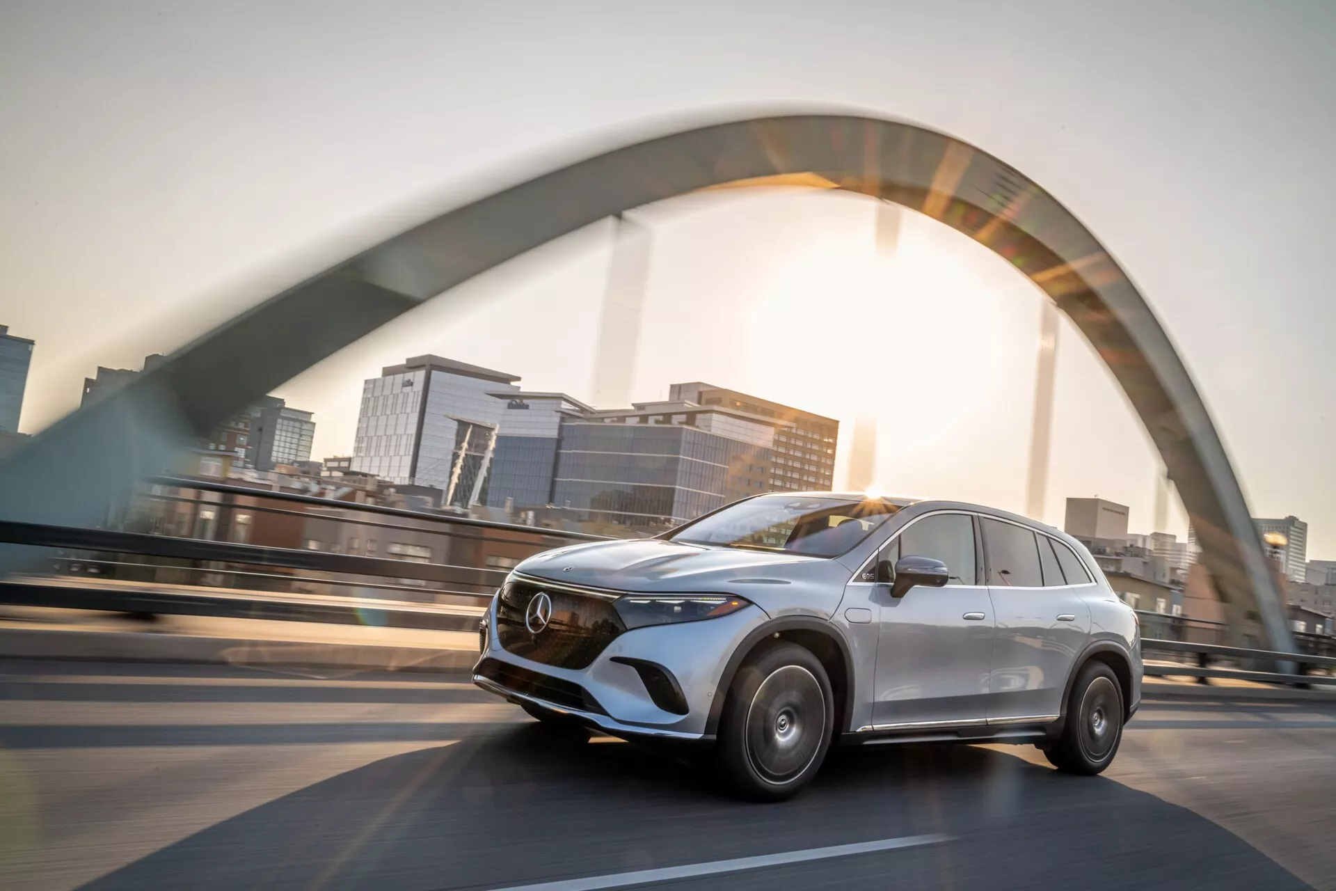 Obrázok zobrazuje strieborný Mercedes EQS-SUV 2022 jazdiaci po zvýšenom moste. Zachytený z nízkeho uhla, predný pohľad z troch štvrtín zdôrazňuje moderný dizajn SUV. Na strane vodiča je viditeľná výrazná chrómová mriežka s emblémom, elegantné línie, štylizované LED svetlomety a rozmazané kolesá, čo naznačuje rýchlosť. Osvetlenie slnečných lúčov a jemný obrys mesta pridávajú dramatický efekt, podčiarkujúc luxus a pokročilé inžinierstvo.