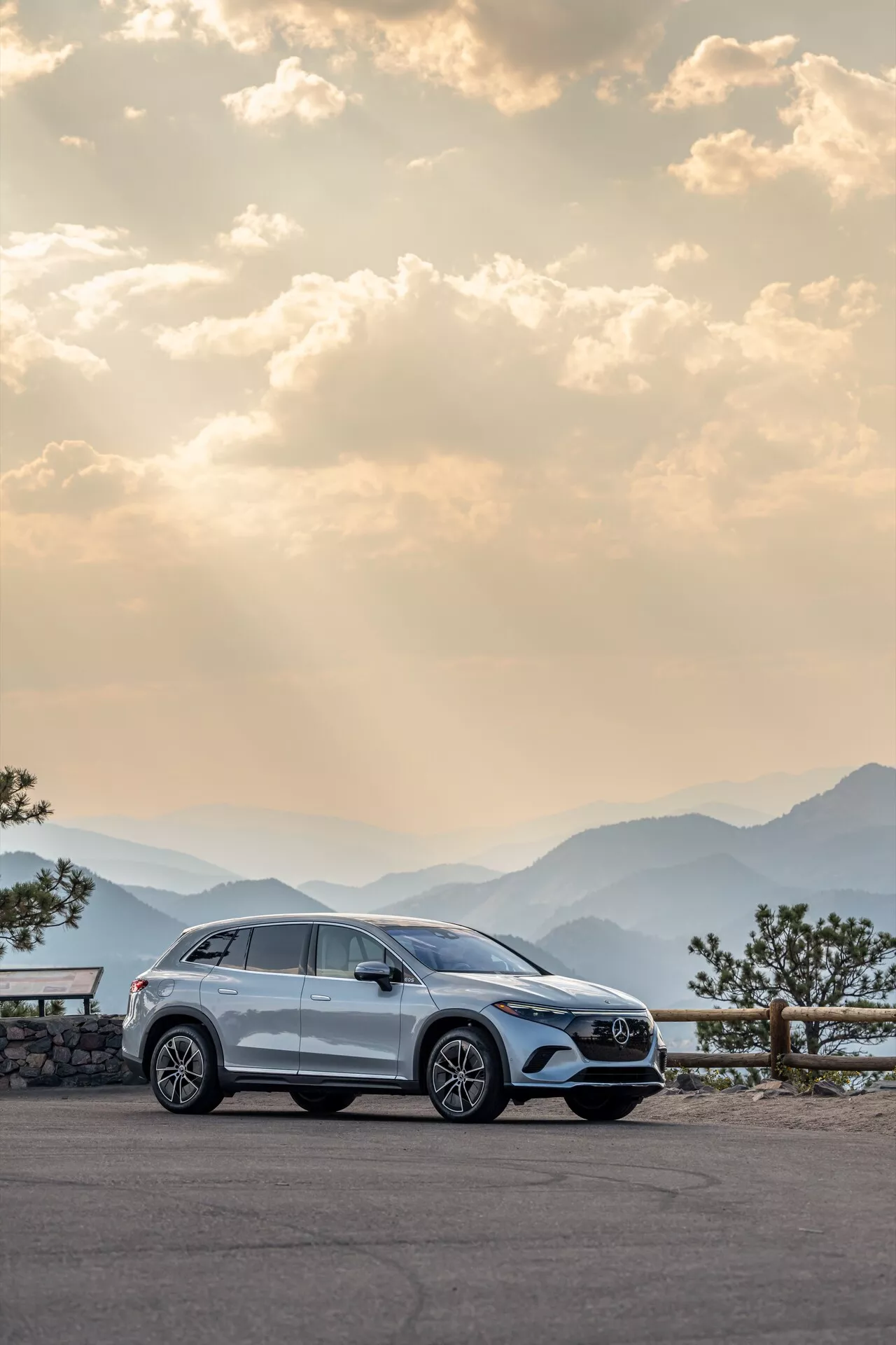Moderný Mercedes EQS-SUV 2022 zaparkovaný na malebnej vyhliadke, zdôrazňujúci svoj pravý bočný profil s hliníkovými kolesami, kľučkami dverí, bočnými zrkadlami, okennými lištami a kolesami mierne natočenými doľava. Fotografia bola zachytená za úsvitu alebo súmraku s teplým, rozptýleným osvetlením, na pozadí panoramatických pohorí a mierne zamračenej oblohy, zdôrazňujúc luxus, dobrodružstvo a vysokú kvalitu remeselného spracovania.