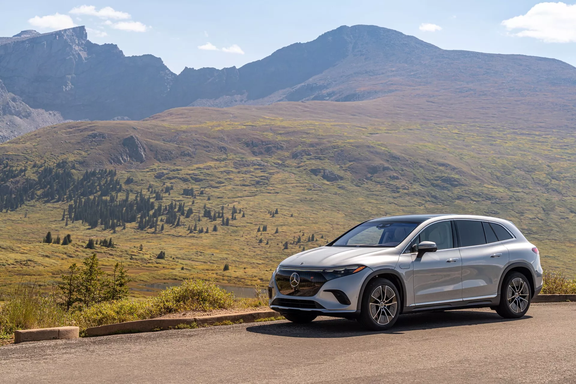 Obrázok zobrazuje moderný Mercedes EQS-SUV 2022 umiestnený na asfaltovej ceste s malebnou horskou scenériou na pozadí pod jasnou oblohou s rozptýlenými mrakmi. Pohľad z troch štvrtín zpredu odhaľuje kľúčové prvky, ako je mriežka chladiča s emblémom, pokročilé svetlomety a dizajn predného nárazníka s čidlami alebo kamerami. Aerodynamické línie na kapote a bokoch spolu s bočným profilom zobrazujúcim zliatinové kolesá, bočné zrkadlá, charakterové línie a sklon strechy zdôrazňujú dizajnové a technologické prvky vozidla.