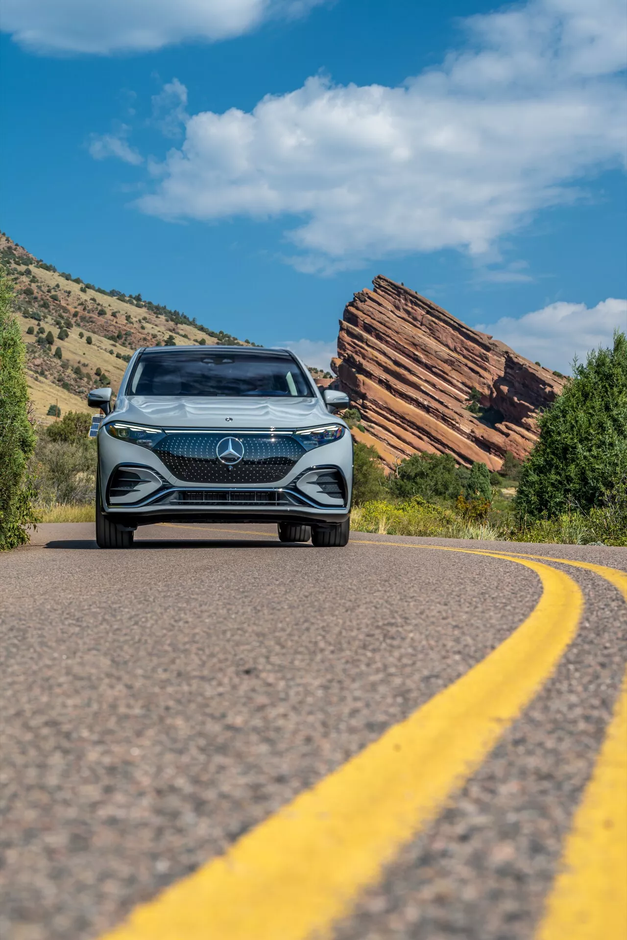 Nízkoúrovňová snímka Mercedes EQS SUV 2022 približujúceho sa na kľukatej ceste, zobrazujúca odvážnu prednú masku s veľkým hviezdnym emblémom, zložitý diamantový dizajn, LED svetlomety a denné svetlá, na pozadí jasnej modrej oblohy a členitého prírodného terénu s vrstvovými skalnými formáciami.