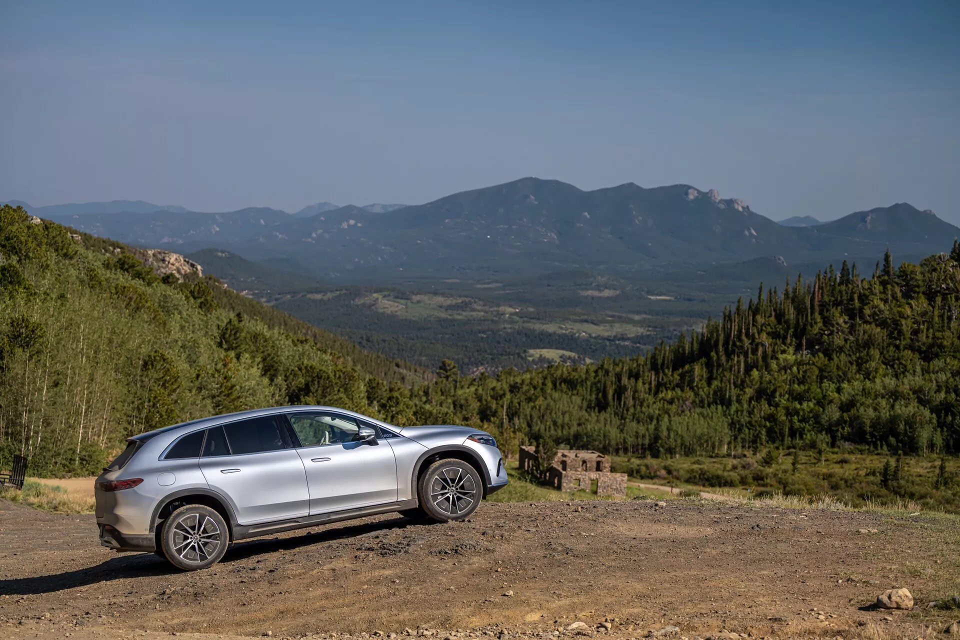 Bočný profil strieborného Mercedesu EQS-SUV 2022 na nerovnom teréne s malebným horským pozadím, zachytený pod uhlom 30 až 40 stupňov. Vlastnosti zahŕňajú vysokú svetlú výšku, zvýšenú výšku jazdy, veľké robustné kolesá, tvarované línie na dverách, možnú panoramatickú strechu, integrované smerovky v bočných zrkadlách, tónované okná a moderné LED svetlomety a koncové svetlá.