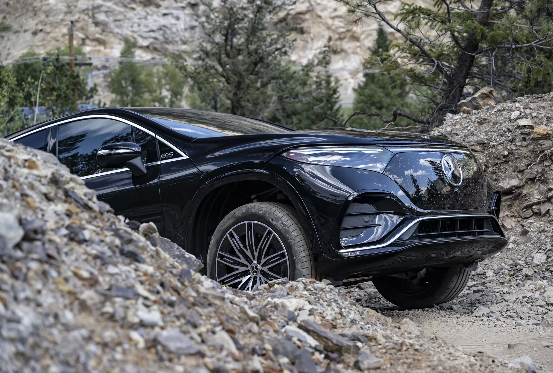 Obrázok čierneho Mercedesu EQS-SUV 2022, ktorý prechádza terénom z nízkeho bočného profilu. Zameranie je na prednú štvrtinu vozidla, s detailmi ako lesklý čierny povrch, elegantné línie, predná mriežka s emblémom, charakteristické svetlomety, svalnaté oblúky kolies, zliatinové kolesá a dezén pneumatík vhodný do každého terénu. Predné koleso je mierne otočené doprava, čo naznačuje manéver. Prírodné pozadie s kameňmi a riedkou vegetáciou zdôrazňuje terénne schopnosti SUV.