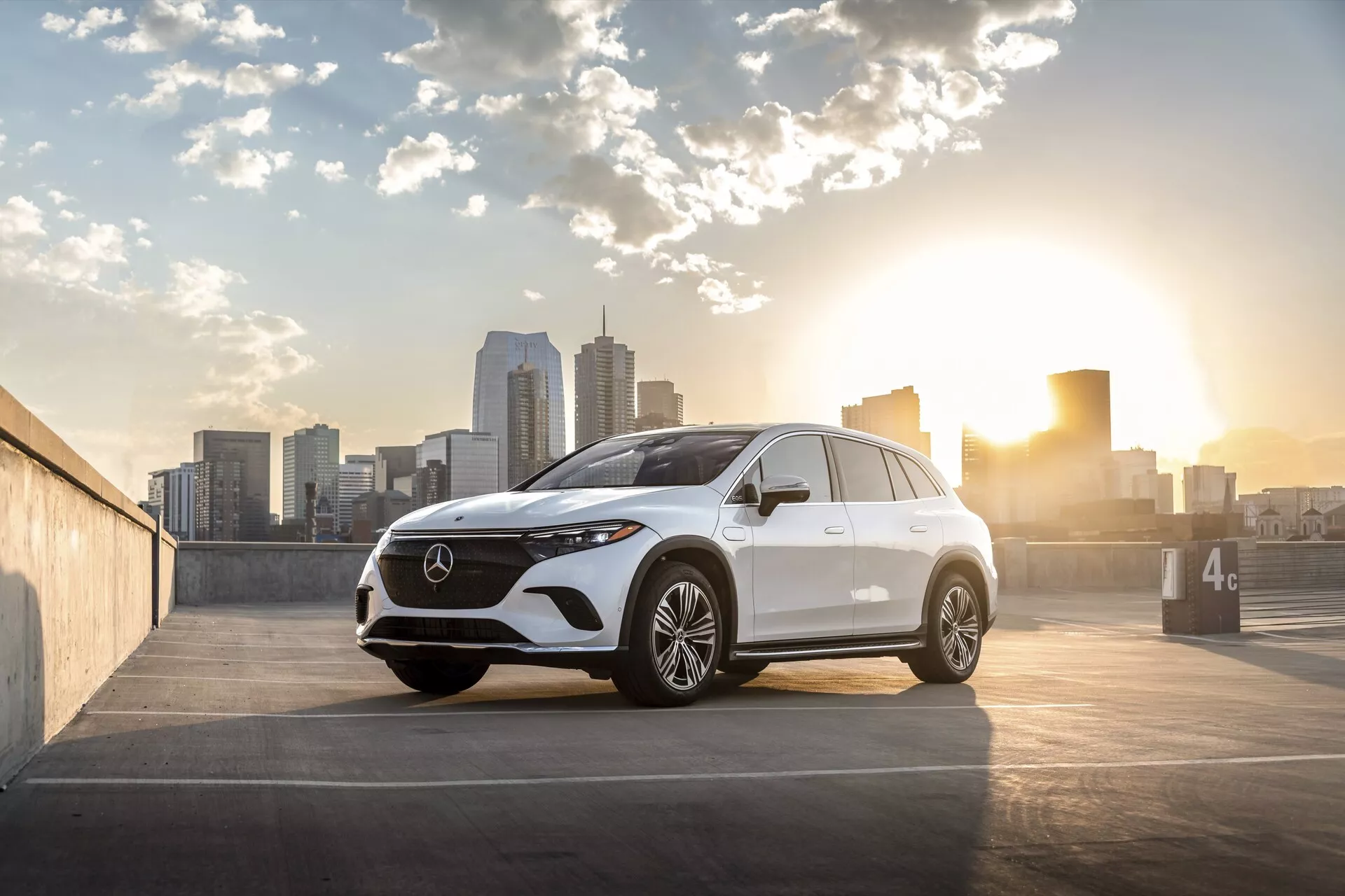 Obrázok ukazuje moderný Mercedes EQS-SUV 2022 umiestnený na vonkajšom parkovisku s panorámou mesta v pozadí, zachytený buď pri východe alebo západe slnka s teplým osvetlením zozadu. Pohľad z troch štvrtín spredu zdôrazňuje mriežku chladiča, svetlomety, dizajn predného nárazníka, kapotu a stranu vodiča, fotografovaný z nízkeho uhla blízko úrovne kolies. Detaily obsahujú prominentnú mriežku s veľkým emblémom, elegantné LED svetlomety, prednú kameru alebo senzorový klastr, viaclúčové zliatinové kolesá, integrované smerovky na bočných zrkadlách, jemné kontúrové línie pre aerodynamiku a mierne klesajúcu líniu strechy. Skladba fotografie umiestňuje auto napravo od rámu s panorámou mesta a slnkom naľavo, pridávajúc odlesk a umeleckú príťažlivosť, vhodné pre kontexty automobilového priemyslu, luxusu a mestského životného štýlu.