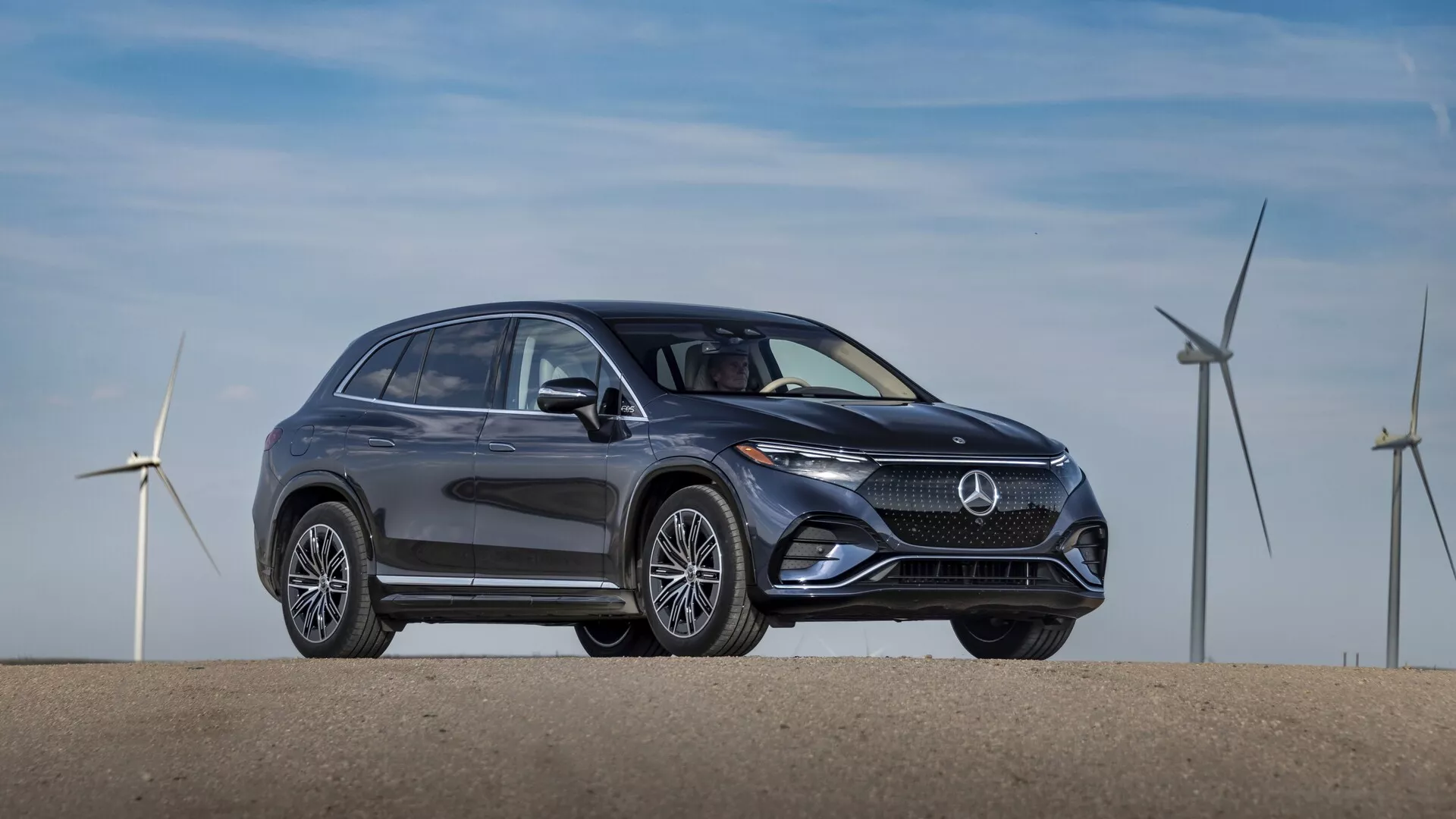 Bočný profil SUV Mercedes EQS 2022 v tmavej farbe, zaparkovaného na rovnom povrchu. Obrázok, zachytený z mierneho predného štvrtinového uhla, ukazuje elegantný a moderný dizajn so zakrivenými líniami a hladkými povrchmi, ktoré zdôrazňujú aerodynamickú účinnosť. Významné prvky zahŕňajú výraznú mriežku s centrálnym logom značky, elegantné LED svetlomety, nasávacie otvory alebo snímače v spodnom nárazníku, sofistikované viaclúčové kolesá z hliníkovej zliatiny, bočné zrkadlá s integrovanými smerovkami, okná s chromovanými rámami a jemne sa zvažujúcu strechu s nosičmi na streche. V pozadí sú veterné turbíny, ktoré naznačujú ekologickú alebo elektrifikovanú povahu vozidla.