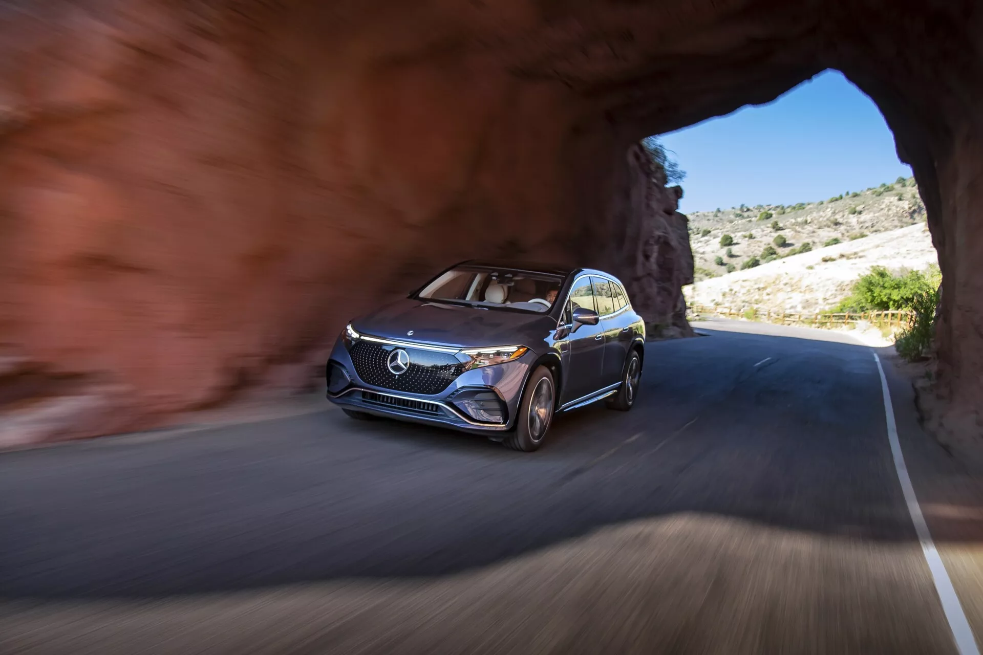 Obrázok auta Mercedes EQS-SUV 2022 vychádzajúceho z tunela na cestu, nasnímaný z nízkeho uhla zameraného na prednú ľavú stranu, ukazuje moderný dizajn vozidla s výraznou mriežkou, logom, dennými LED svetlami, viacklučovými zliatinovými diskmi a kovovým lakom. Auto je v pohybe, čo naznačujú rozmazané pozadie a kolesá. Okolie ukazuje skalnatý terén s riedkou vegetáciou, čo naznačuje horskú alebo púštnu oblasť za jasného počasia.