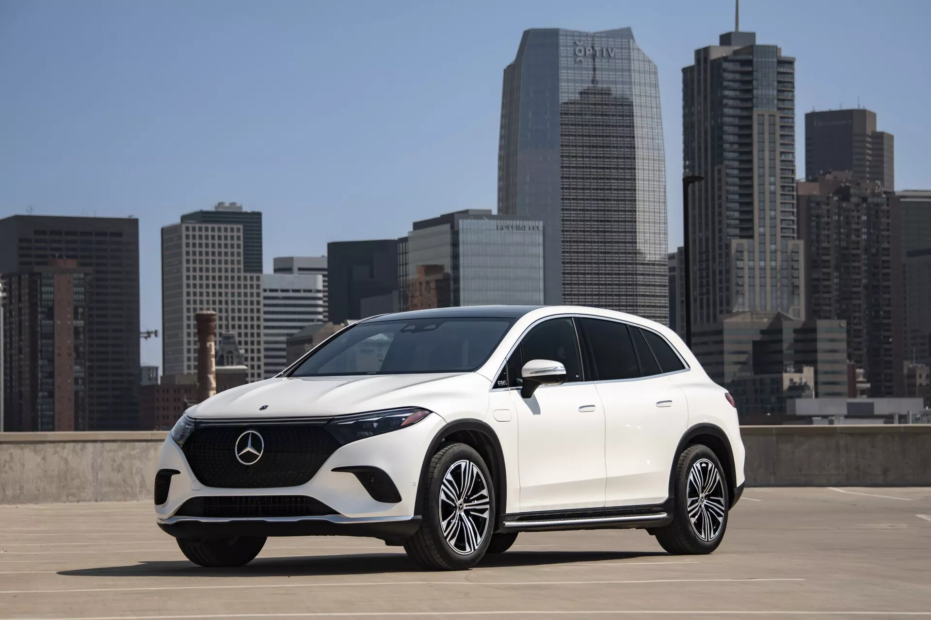Trojštvrtinový predný pohľad na biely Mercedes-Benz EQS SUV z roku 2022 na hornom poschodí parkovacej stavby s mestským panorámom v pozadí. SUV má elegantný dizajn s hladkými líniami, veľkú prednú masku s emblémom Mercedes-Benz a LED svetlomety. Predný nárazník má veľké vstupy vzduchu a strieborný kryt, a viacpólové zliatinové kolesá dodávajú dotyk luxusu. Pokročilá technológia zahŕňa možné senzory alebo kamery v prednej maske, integrované smerovky v bočných zrkadlách a aerodynamický dizajn naznačuje úsporu paliva. Slnečné svetlo vrhá tiene smerom ku kamere, čo zvýrazňuje dizajn vozidla.