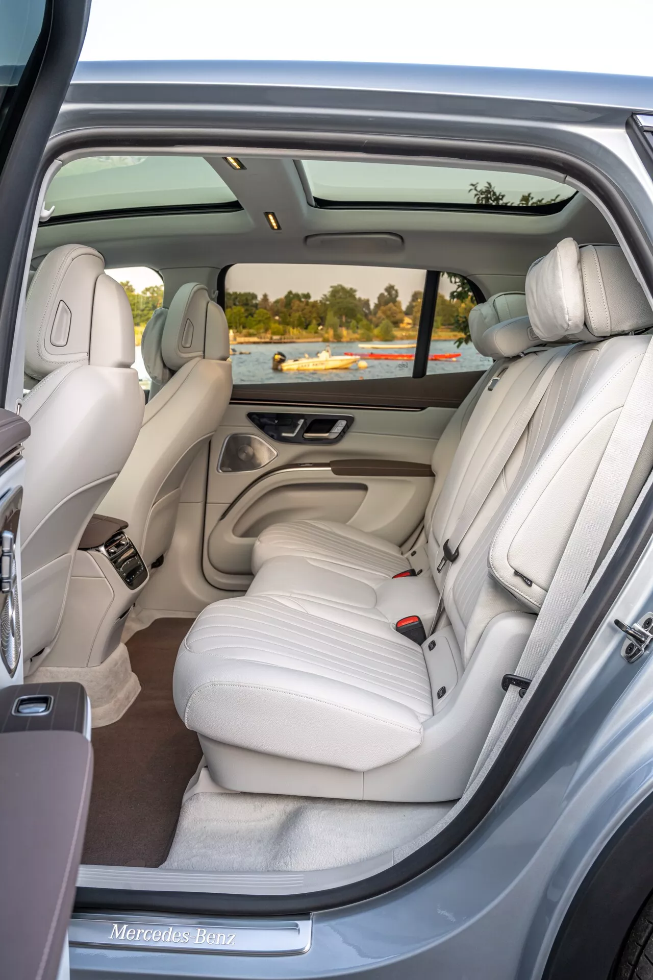 Pohľad na interiér Mercedesu EQS SUV 2022 z pravých zadných dverí, ktorý ukazuje luxusnú zadnú lavicu z krémovej kože so tromi opierkami hlavy, panoramatickú strechu, elektronické ovládacie prvky, individuálne klimatizačné ventilačné otvory, chrómové akcenty, mriežku reproduktora, úložnú kapsu a vysokokvalitný koberec.