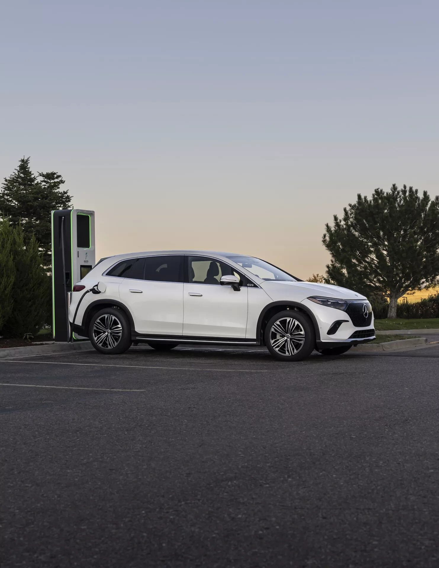 Moderný Mercedes EQS-SUV 2022 pri nabíjacej stanici za súmraku, fotografovaný z bočného-predného uhla, zvýrazňujúci jeho elegantný profil, zliatinové kolesá a nabíjací port.