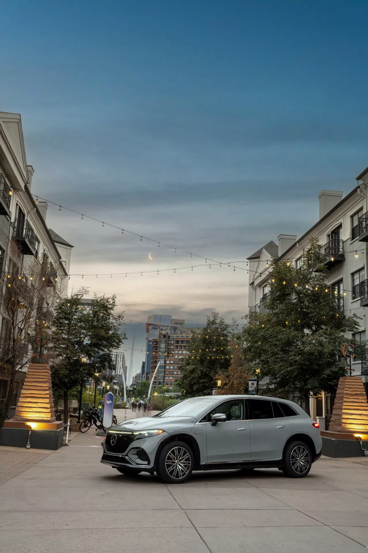 Predný trojštvrťový pohľad na strieborný Mercedes EQS-SUV 2022, zdôrazňujúci jeho výraznú mriežku s emblémom, LED denné svetlá, zliatinové kolesá a štýl karosérie. Auto je zaparkované v luxusnom mestskom prostredí s dekoratívnym osvetlením a budovami, zdôrazňujúc jeho luxusný a moderný dizajn.