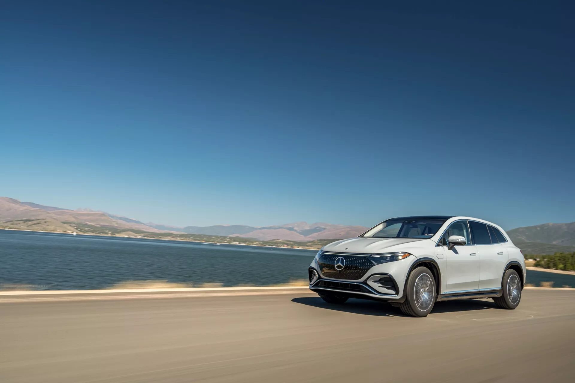 Moderný luxusný crossover SUV, Mercedes-Benz EQS, jazdiaci po asfaltovej ceste blízko veľkého vodného telesa. Obrázok zachytáva auto z troch štvrtinového čelného pohľadu za jasného denného svetla a zvýrazňuje prvky ako LED svetlomety, charakteristickú masku, aerodynamický dizajn a veľké zliatinové kolesá. Vozidlo je v pohybe, čo naznačuje dynamiku, s jazerom alebo riekou v pozadí a čistou oblohou, čo je typické pre prémiové elektrické modely.
