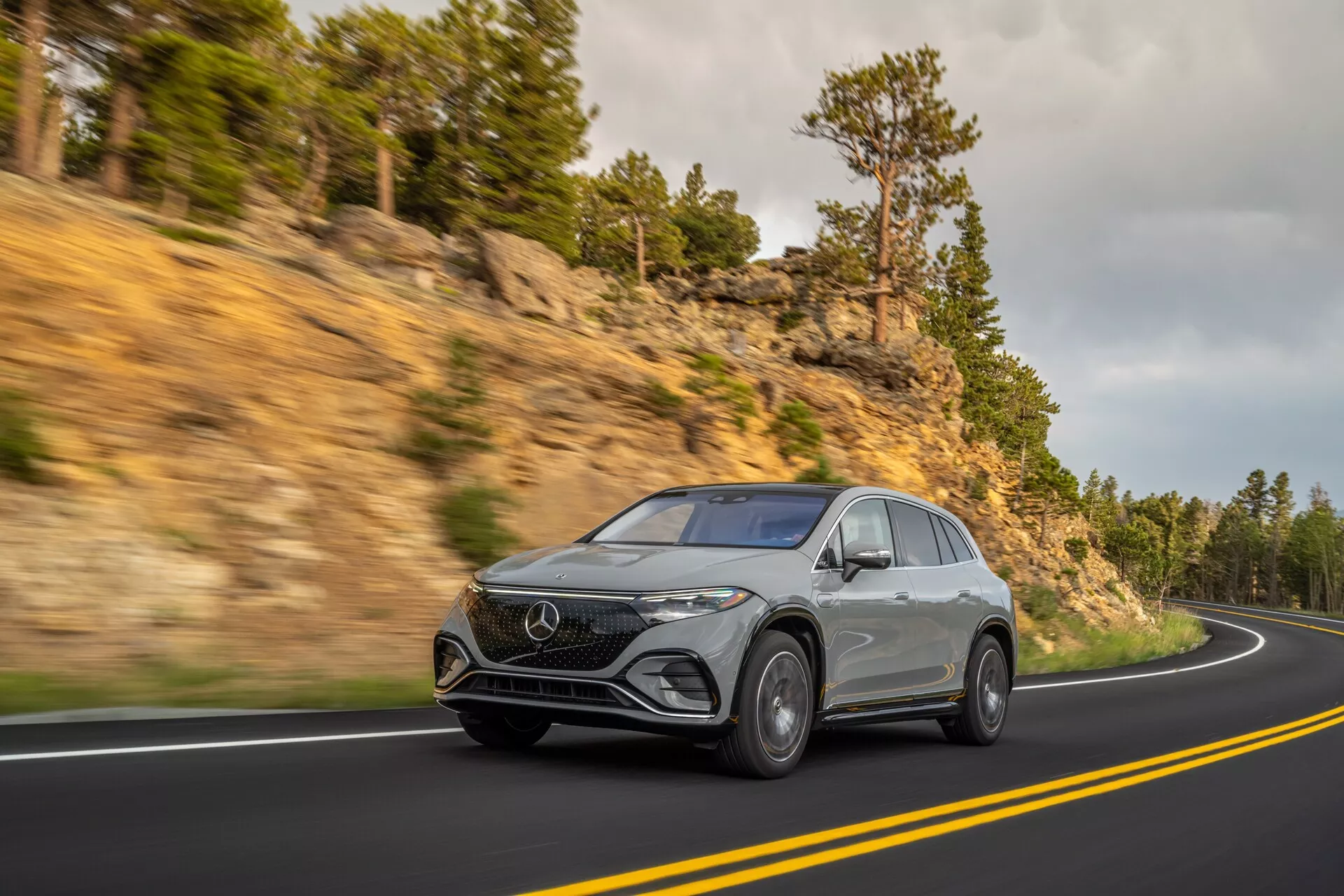 Dynamický záber strieborného Mercedes EQS SUV 2022 na kľukatej horskej ceste, pohľad z trojštvrťového predného uhla, aby vynikla jeho výška a prítomnosť na ceste. S modernými LED svetlometmi, výrazným predným logom, luxusnými zliatinovými kolesami a aerodynamickými líniami potvrdenými súčasným dizajnom.