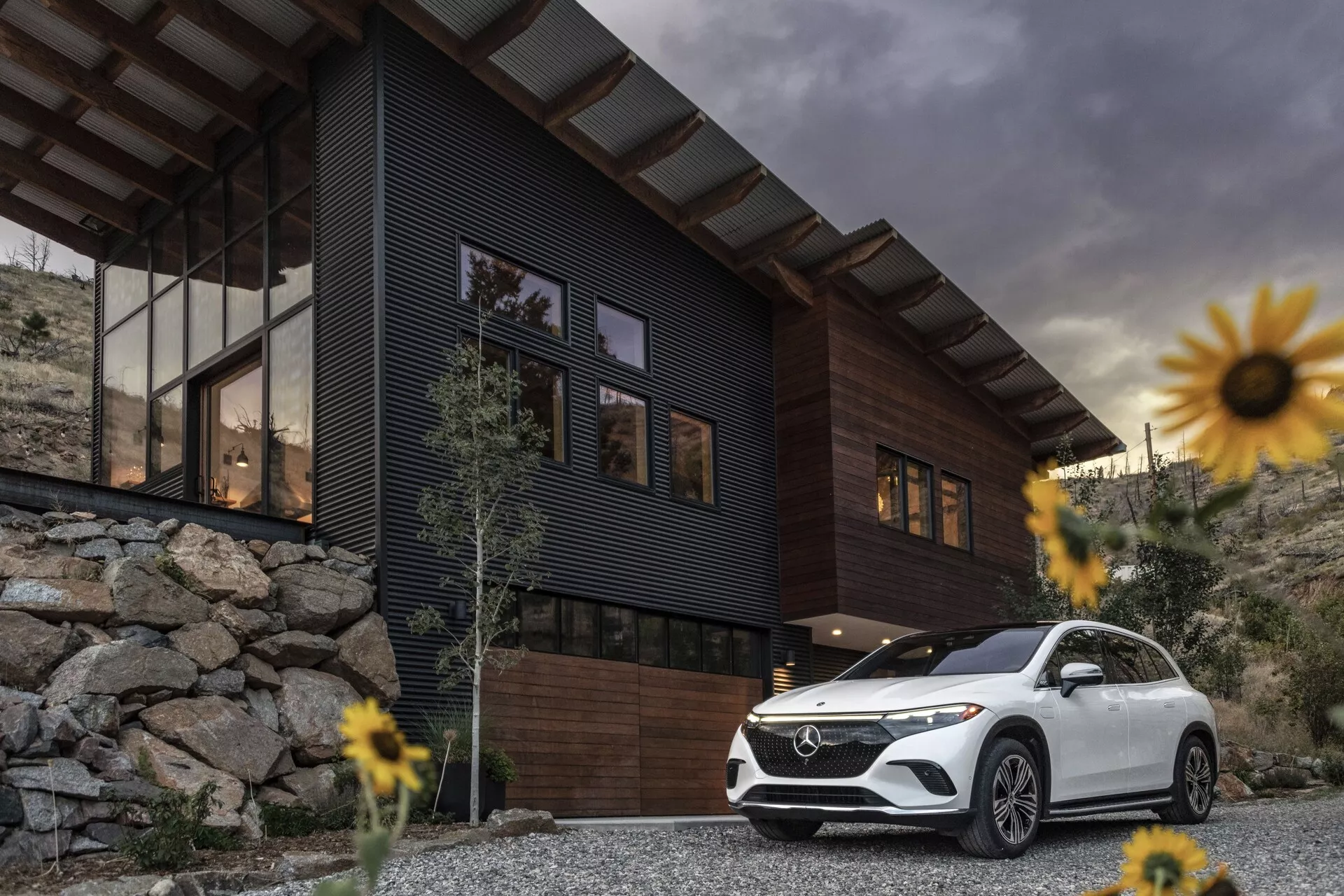 Obrázok luxusného SUV Mercedes-Benz EQS 2022 zaparkovaného pred moderným domom s oblačnou oblohou v pozadí. Záber z nízkeho uhla zdôrazňuje postoj a veľkosť vozidla, zameriava sa na prednú masku, zostavu svetlometov, predný nárazník, kontúry kapoty a ľavú stranu vodiča až po zadné dvere. Medzi prvky patria LED denné svetlá a elegantné kolesá, čo prispieva k pocitu luxusu a pokročilej technológie. Biele SUV kontrastuje s tmavým domom a skalnatou krajinou, obklopené mierne rozmazanými kvetmi vďaka malej hĺbke ostrosti, ktorá zvýrazňuje SUV ako hlavný bod.