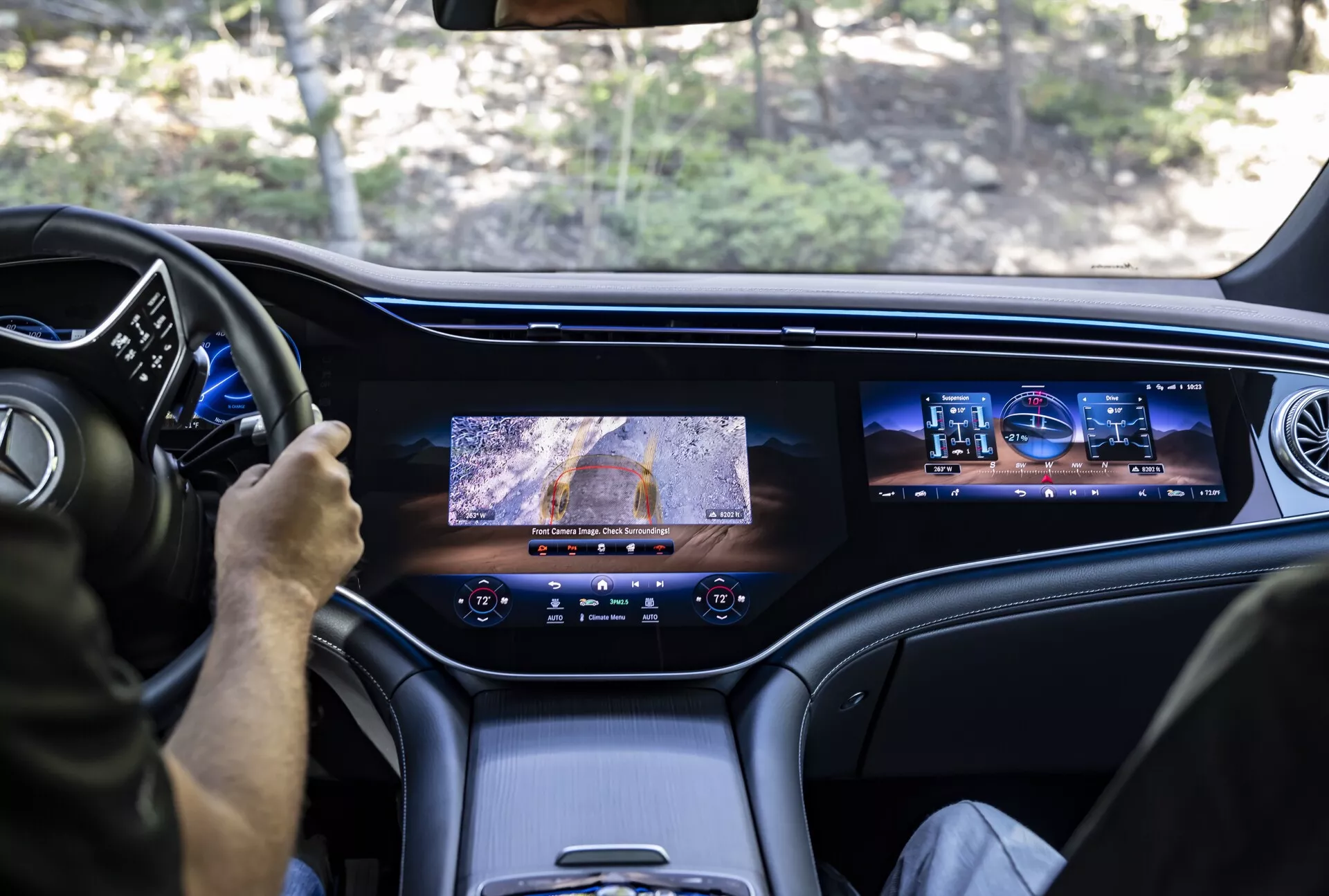 Pohľad zo zadného sedadla Mercedesu EQS-SUV 2022 ukazujúci prednú časť interiéru; zobrazuje vodiča používajúceho multifunkčný volant, digitálny prístrojový panel, veľkú centrálnu obrazovku s kamerovým výhľadom, sekundárnu infotainmentovú obrazovku pre spolujazdca, prémiové kožené alebo vysoko kvalitné materiály interiéru a moderné dizajnérske ventilačné otvory, zdôrazňujúce luxusné a pokročilé technologické vlastnosti vozidla.