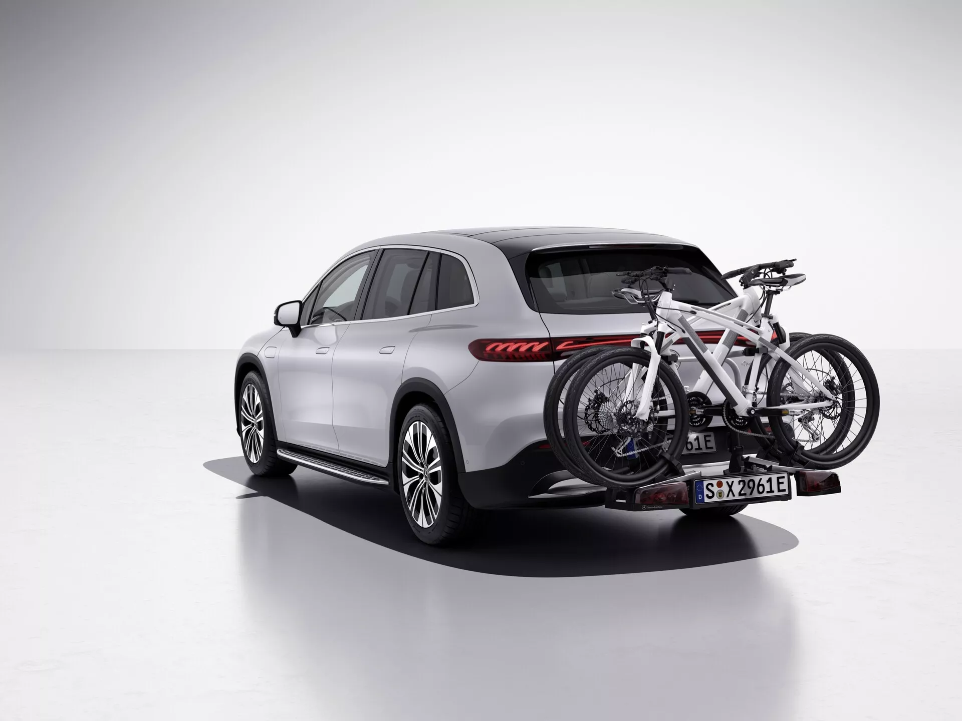 Obrázok zobrazuje SUV Mercedes EQS z roku 2022 v štúdiovom prostredí s čistým, bielym pozadím, zobrazený z zadného trojštvrtinového uhla s pohľadom na stranou pre pasažierov. Vozidlo je vybavené nosičom bicyklov na zadnom ťažnom zariadení, ktorý drží jeden bicykel. SUV sa vyznačuje elegantným, moderným dizajnom s hladkými líniami, výrazným zadným svetelným klastrom s horizontálnymi červenými LED zadnými svetlami, viaclúčovými zliatinovými diskami a karosériou natretou v svetlej farbe, pravdepodobne striebornej alebo sivej, v kontraste s čiernymi spodnými ozdobami a obkladmi oblúkov kolies. Bočné zrkadlá, strešné ližiny a obruby okien sú v tmavšom tóne, s panoramatickou strechou, ktorá pridáva prémiový pocit. Tento obrázok zdôrazňuje prvky ako LED osvetlenie, športový dizajn kolies a bicyklový nosič, čo ho robí vhodným pre kategórie ako Vozidlá, Automobilová technológia, Športové/vonkajšie vybavenie a luxusné SUV.